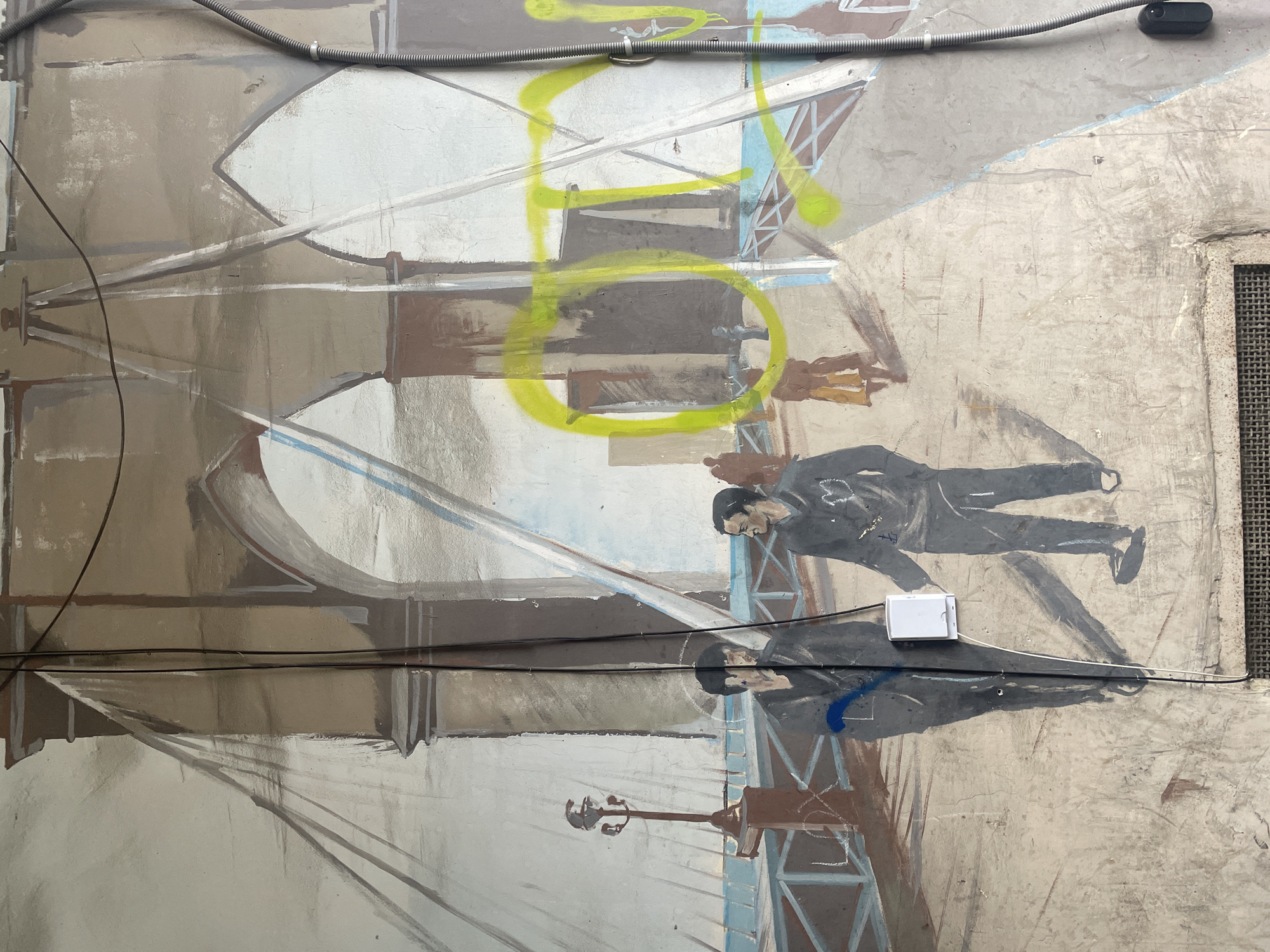 A vertical photograph captures a section of a weathered mural on a wall in Yerevan, Armenia. The mural depicts a stylized representation of a suspension bridge, identified by its brown stone towers featuring arched passageways and numerous cables extending over a blue body of water. Two male figures, dressed in dark clothing, are prominent in the foreground, walking on the bridge deck. The figure on the left is partially obscured by an affixed white electrical junction box and black cables. The figure on the right displays a light-colored heart emblem and an anchor symbol on his chest. Additional indistinct figures are painted further back on the bridge, including one in a light brown coat. A single streetlamp with a dark pole and light fixture is depicted on the left side of the bridge deck.
The mural surface shows significant signs of wear, including cracks and discoloration. Bright yellow spray paint graffiti is extensively applied across the central and right portions of the mural, forming large circular shapes and indistinct letters. Blue spray paint appears as a splatter on the left foreground figure and as a word resembling "Love" or "Lox" on a painted bridge railing. Multiple black electrical cables are secured vertically over the mural, partially obscuring painted elements and figures. A white rectangular electrical junction box is mounted on the wall over the lower-left section of the mural. A grey flexible conduit runs vertically along the far right edge of the wall. A rectangular metal grate is visible at the bottom center of the frame.