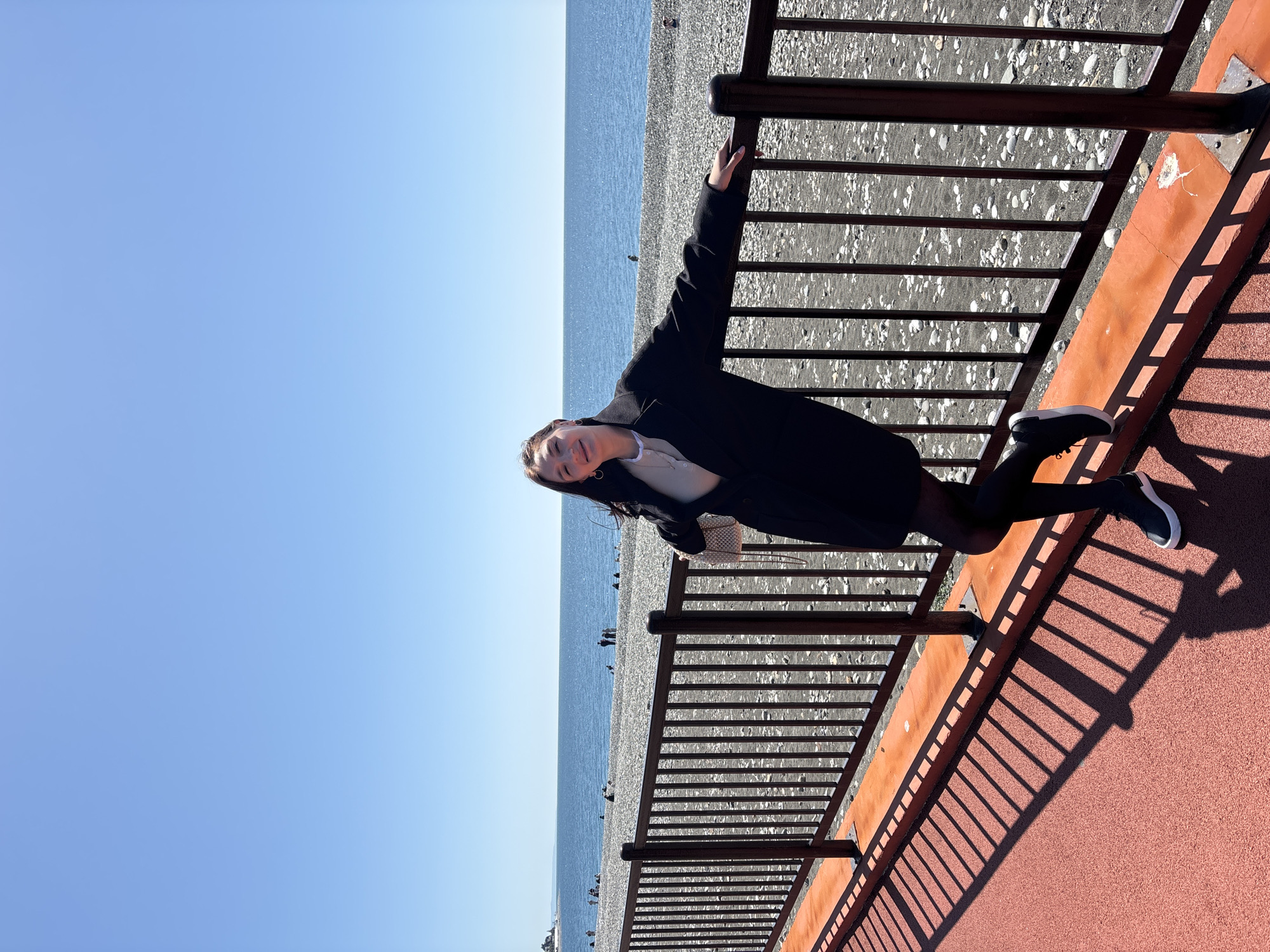 A young woman is prominently featured, standing on a promenade and facing the camera with a gentle smile. She is dressed in a dark, long-sleeved coat or jacket over a light top, black tights, and black and white sneakers. Her left arm rests casually on a dark metal railing that separates the promenade from the beach.
The scene depicts a sunny coastal environment. The promenade has a distinctive reddish-orange paved surface, upon which the railing casts long, sharp shadows, indicating a bright day, likely mid-morning or afternoon. Beyond the railing lies a wide pebble beach, dotted with several small, indistinct figures of other people. The calm blue sea stretches out to the horizon, meeting a clear, light blue sky.
The overall mood is tranquil and pleasant, with the woman appearing relaxed and content. The setting, characterized by a pebble beach, calm sea, and a promenade overlooking the water, aligns with the context of Sochi, Russia. No visible text is present in the image.