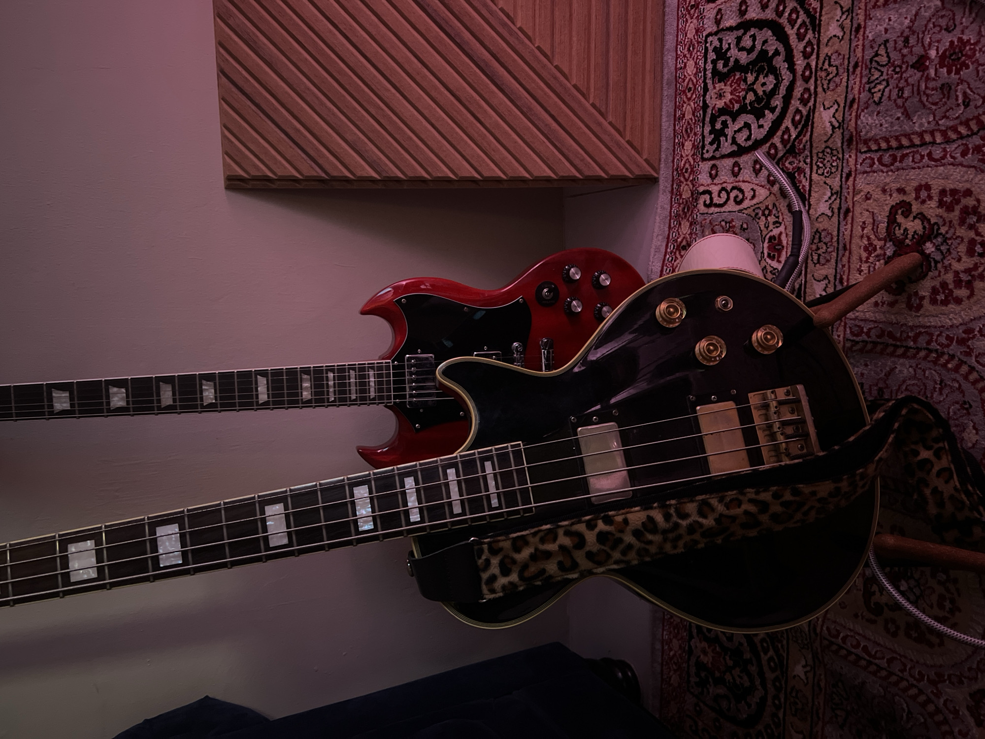 The image shows two electric guitars, one black and one red, leaning against a wall. The black guitar is in the foreground and has a leopard-print strap. The red guitar is in the background and has a body shape similar to an SG model. A textured wooden acoustic panel is propped up against the wall to the right of the guitars. The guitars are placed on a patterned rug, and a cable runs from the black guitar to the floor. The lighting in the scene is dim and has a pinkish hue, possibly from stage lighting. The location is not specified, but the image is described as being from Singapore.
