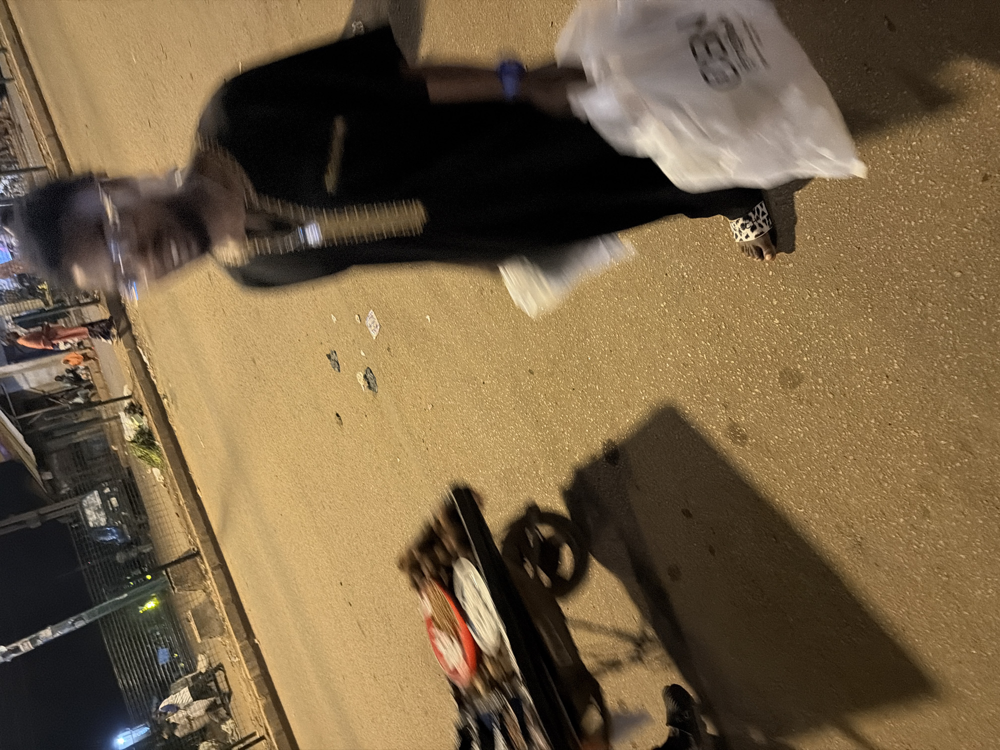 A person, possibly male based on attire, walks on a paved street at night in Bukuru, Nigeria, holding two white plastic bags. The individual is wearing a dark garment with gold embellishments around the neck and a patterned sandal on their right foot. Their face is blurred, suggesting motion. In the background, a food cart with items on display, a vehicle, and other people are visible. The street appears to be lit by artificial light, and there is some debris on the ground.
