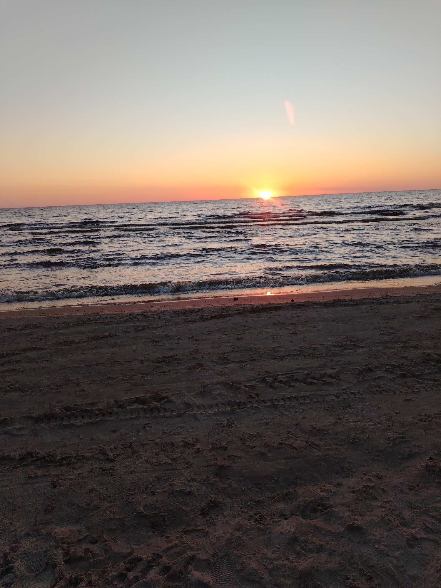 The image depicts a serene beach scene at sunset. The sky transitions from a pale blue at the top to warm orange and yellow hues near the horizon. The sun is low in the sky, casting a radiant glow over the rippling water. Gentle waves lap at the sandy shore, creating a peaceful atmosphere. The sand itself appears undisturbed, with faint tire tracks visible, suggesting recent activity. The overall impression is one of tranquility and natural beauty. This location is Ivanovskoe, Russia.