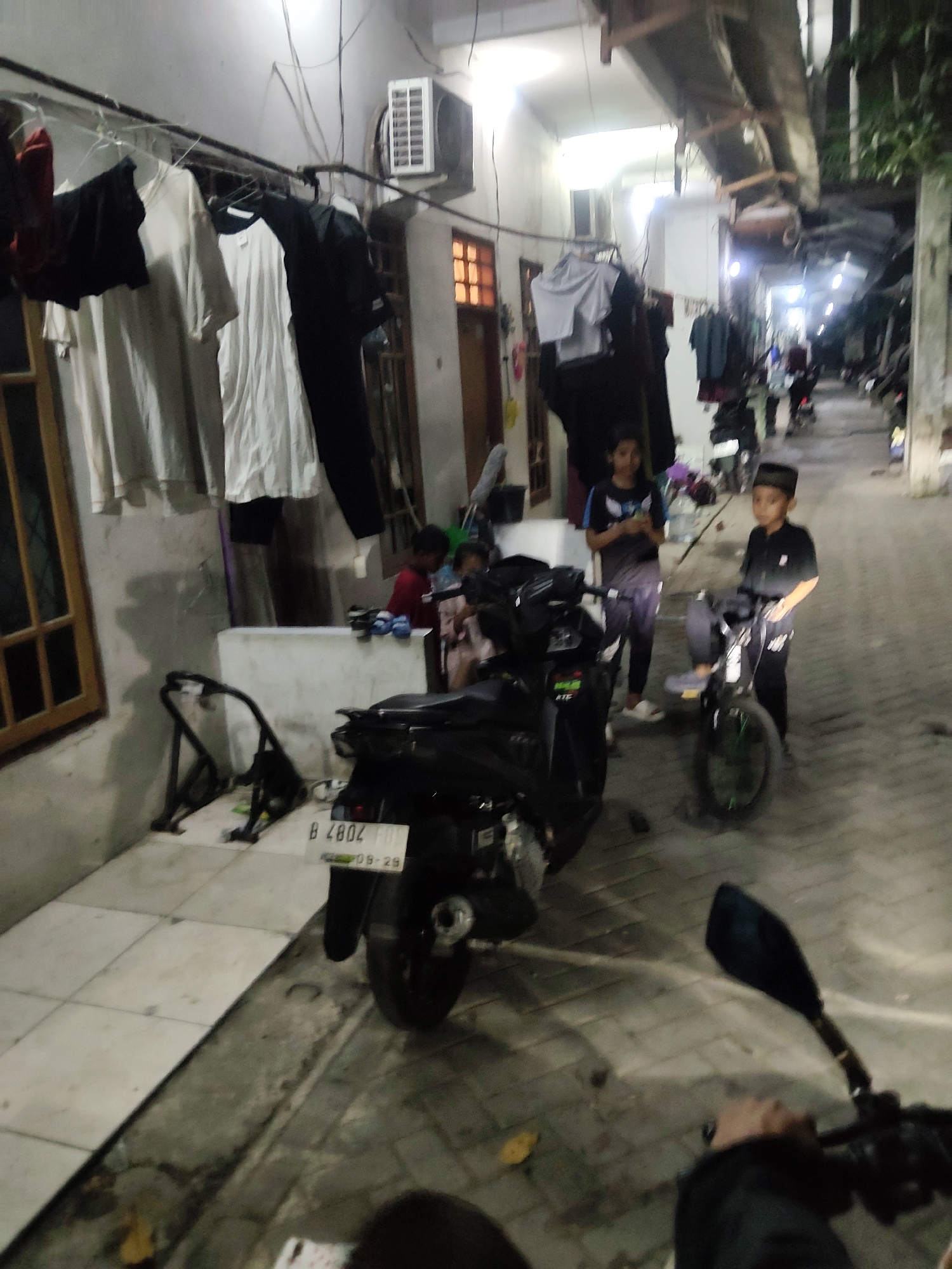In Mekarmukti, Indonesia, an outdoor alleyway is depicted at night. Clothes are hanging on a clothesline stretched across the scene, with several garments including white t-shirts and dark fabrics visible. Two young boys are present; one is on a bicycle with green wheels, wearing a dark shirt and a peci hat, while the other stands nearby. A motorcycle is parked in the foreground, its license plate reading "B 4802 FUK" with a date "09-28". Another motorcycle is visible further down the alleyway with a rider. The alley is lined with buildings, one of which has a window with orange panes. An air conditioning unit is mounted on the exterior wall of one building. The ground is paved with tiles and concrete. The scene appears to be a residential or densely populated area.