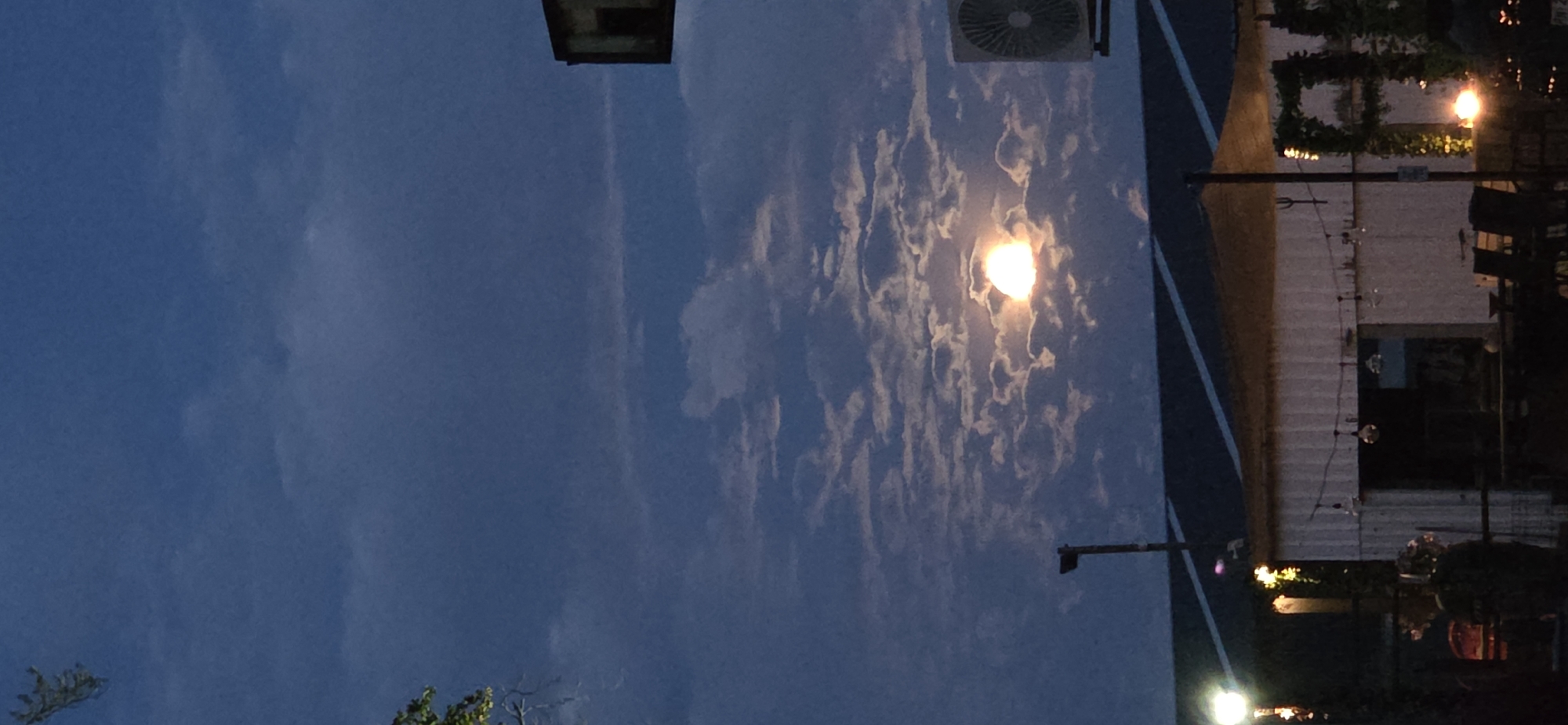 The image shows a nighttime scene at what appears to be an outdoor seating area, possibly a patio or beer garden. The setting is Eeklo, Belgium. The sky is a deep blue, indicating it is nighttime. There are scattered clouds, and a bright, luminous full moon is visible, partially obscured by the clouds. The moon's glow illuminates the clouds around it.
In the foreground, there are some trees with green foliage, suggesting a natural element to the location. To the right, there is a building with a white, corrugated facade. String lights with small, clear bulbs are hung along the building, and some of these lights are illuminated, casting a warm glow. A street lamp is also visible, emitting a bright yellow light. There are tables and chairs visible in the background, suggesting a place where people gather.
Above the building, an air conditioning unit is mounted on the wall. There is also a dark, rectangular structure on a pole, possibly part of a light fixture or signage. The overall mood of the image is peaceful and serene, with the soft light of the moon and the string lights creating a gentle ambiance. The scene suggests a calm evening, perhaps after sunset. There is no visible text in the image.
