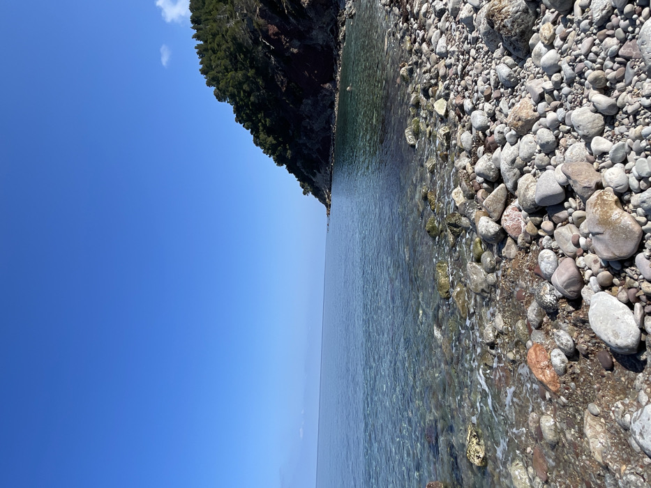 This image displays a coastal scene, specifically a rocky beach located near Valldemossa, Spain. The foreground features a wide expanse of beach composed of smooth, rounded pebbles and stones of varying sizes and earthy tones, including grey, brown, and reddish hues. The clear, calm ocean water, exhibiting shades of turquoise and blue, gently laps the shore, with the rocky seabed visible through its transparent surface. Small white foam lines indicate minimal wave activity along the water's edge.
To the right, a steep hillside ascends, densely covered with green coniferous trees, primarily pine, against a backdrop of reddish-brown exposed earth and rock. The coastline extends into the distance on both the left and right, forming a natural bay or cove. The sky above is a vast, unobstructed expanse of bright blue, with a few faint, wispy white clouds barely visible at the extreme upper right. No individuals, animals, or man-made structures are present within the frame. The scene is illuminated by direct sunlight, casting subtle shadows on the hillside.