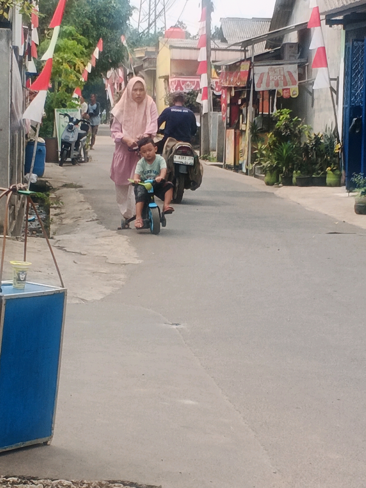In the city of Kadu, Indonesia, a child rides a small blue toy motorcycle down a paved street, followed closely by a woman in a pale pink hijab and matching dress. The child is wearing a light blue t-shirt and dark shorts. Behind them, a person on a motorcycle wearing a dark shirt and jeans drives away. Red and white flags are strung along poles on the left side of the street, and more flags are visible on the right. Buildings line the street, some with businesses displaying signs. Potted plants are arranged along the side of the road. In the foreground, a blue and silver stand with a yellow cup on top is visible. The sky appears overcast.