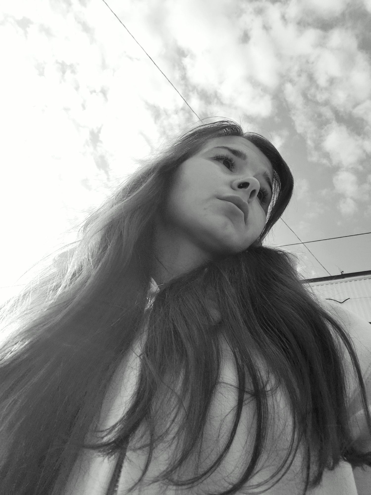 A black and white, low-angle shot shows a woman looking upwards, seemingly pensive, against a backdrop of cloudy sky. A thin wire bisects the upper part of the frame. The woman has long, dark hair that falls around her shoulders and chest. A partial view of a building, possibly with a weather vane, is seen at the upper right. There is nothing to suggest the location is Фоки, Russia.