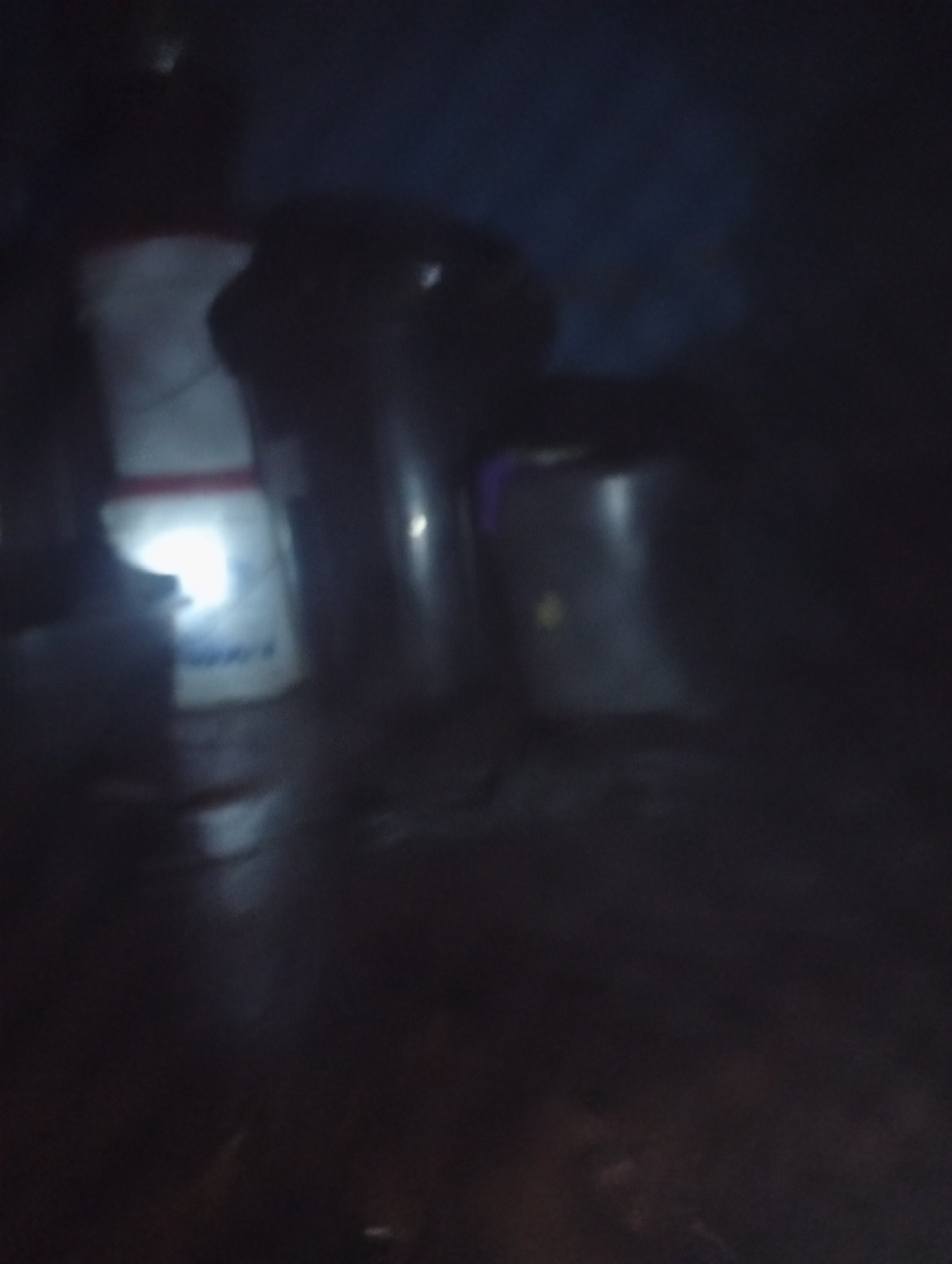 The image is a dark, blurry, and low-resolution photograph that appears to have been taken at night. In the foreground, a large white object with red and blue markings is visible. This object is illuminated by a bright light source, likely a flash. To the right of the white object, there are two large, cylindrical metallic objects. The rest of the scene is obscured by darkness and blurriness, making it difficult to discern any further details about the environment or any other potential subjects or objects. The scene seems to be an indoor or outdoor space with low ambient light conditions.