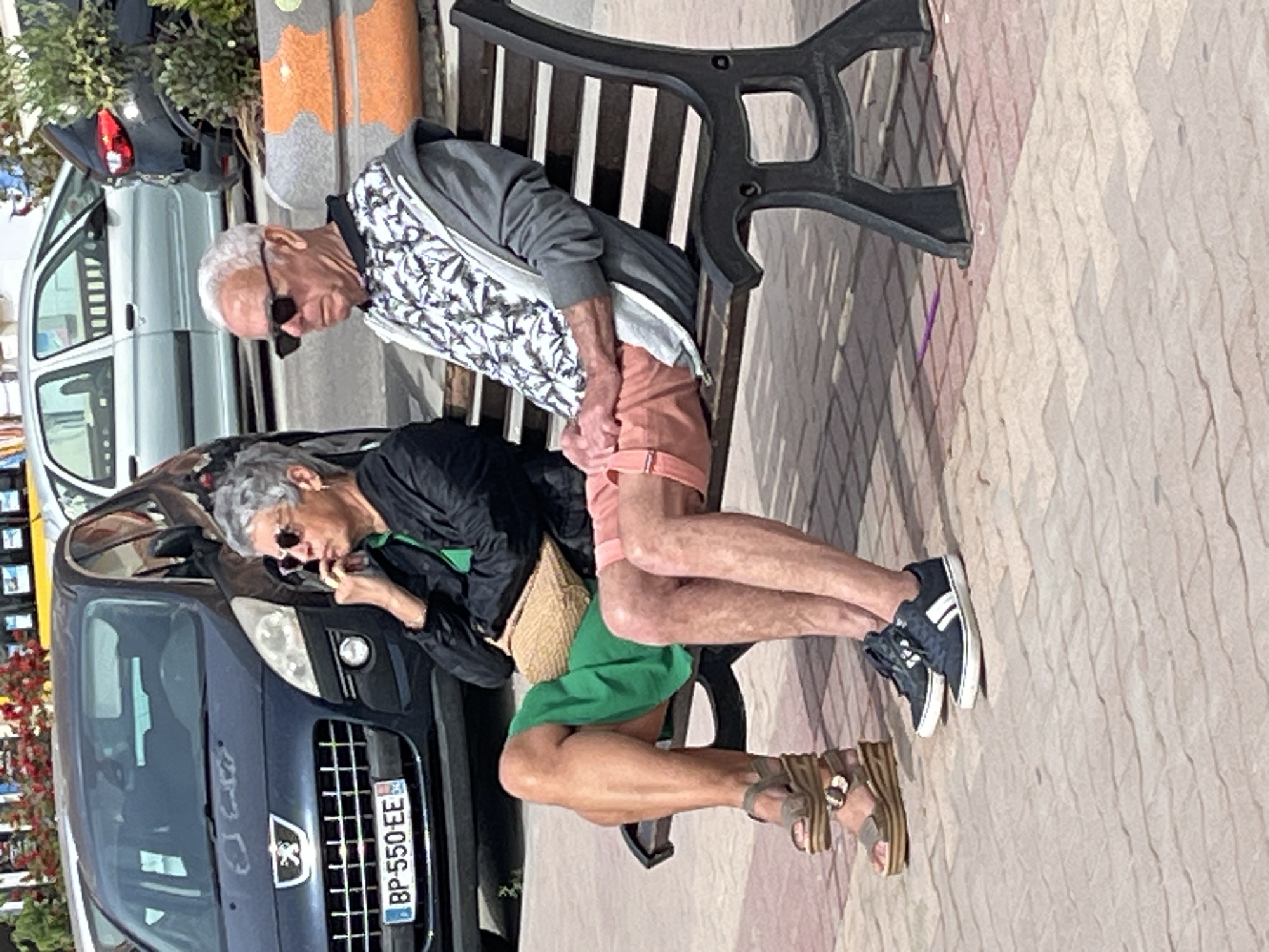An eye-level, full, outdoor shot depicts two light-skinned, older adults sitting on a park bench, next to two parked cars, in Palavas-les-Flots, France. The man, who is on the left side of the frame, is seated on the edge of the bench, with his legs draped over the other person’s legs. He wears a white and grey patterned jacket, pink shorts, and blue and white tennis shoes. The woman sits next to a blue car, wearing a black jacket, a green dress, and tan sandals with thick straps. She is looking down at her hands. The number plate on the blue car reads "BP-550-EE." The setting appears to be a town square near a street, as evidenced by the other parked vehicles and trees. The ground is covered in textured light-colored stone. The photograph is slightly rotated clockwise.