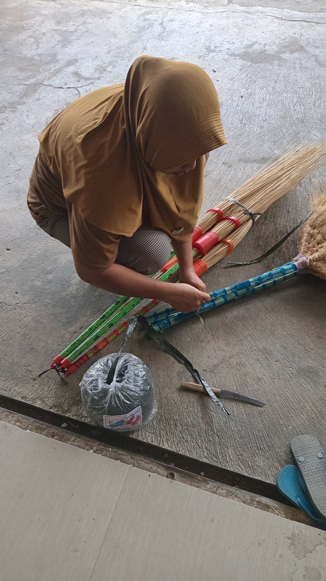 A person wearing a mustard-colored hijab and plaid pants is crouching on a concrete floor in what appears to be an outdoor area. The person is assembling or preparing brooms. In front of the person are several broom heads, some with red and orange bindings, and others with colorful patterned handles. A roll of black string is also visible, as is a small knife with a wooden handle. To the right, a partially visible woven broom head and a pair of flip-flops (one blue, one gray) are on the ground. The concrete floor shows some cracks and weathering. The context suggests this is a scene in Kunduran, Indonesia, likely a residential or workshop area given the materials and tools present.