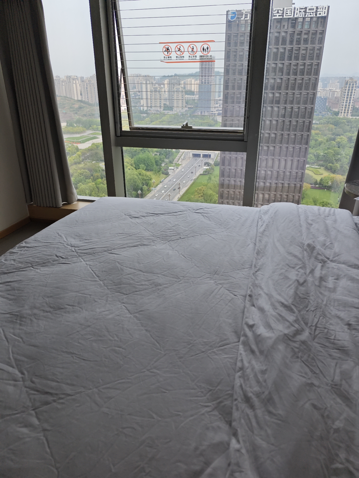 The photo presents a view from inside a room, predominantly featuring a bed covered in a gray wrinkled sheet. Beyond the bed, a large window occupies the center of the frame, partially open to reveal a cityscape. The window has a sign that reads "禁止攀禁止抛物禁止倚靠请看管好您的小孩" which translates to "Do not climb, do not throw objects, do not lean, please take care of your children." The window also has the text "万" and "空国际总部", which could be part of a building name. A gray curtain hangs to the left of the window.
Through the window, a dense urban environment unfolds, including numerous high-rise buildings interspersed with green spaces, suggesting a park or garden landscape. A highway runs through the scene, visible from the window, with cars traveling on it. The architecture of the buildings suggests a modern design, and their arrangement indicates an urban setting, likely in a bustling city such as Liangjiang, China. The light implies a cloudy or overcast day.