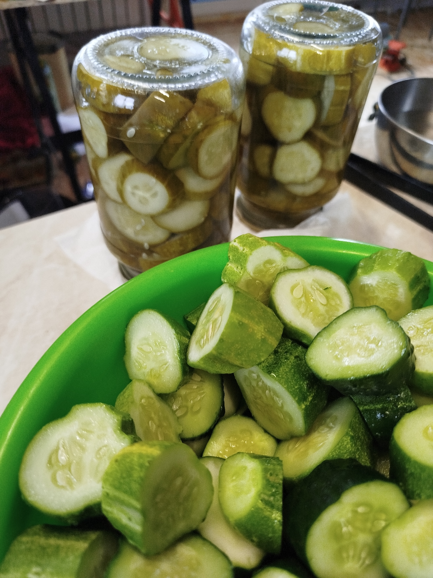 This is a close-up, eye-level shot of sliced pickles in a bright green bowl, positioned in the foreground. The bowl is filled with numerous pickle slices, some whole and some cut in half, revealing their seedy interiors. The pickles appear fresh and crisp. In the background, out of focus, are two glass jars filled with more pickled cucumbers submerged in brine. The jars are cylindrical and topped with clear glass lids. A stainless steel bowl is visible to the right of the jars. The scene appears to be in a kitchen or food preparation area, with a textured tabletop and a hint of other objects and furniture in the blurred background. The lighting is natural and diffused, highlighting the vibrant green of the fresh pickles and the yellowish-green brine in the jars. There are no people visible in the image. The location of Zavodoukovsk, Russia, is not discernable from the visual information provided in the image.