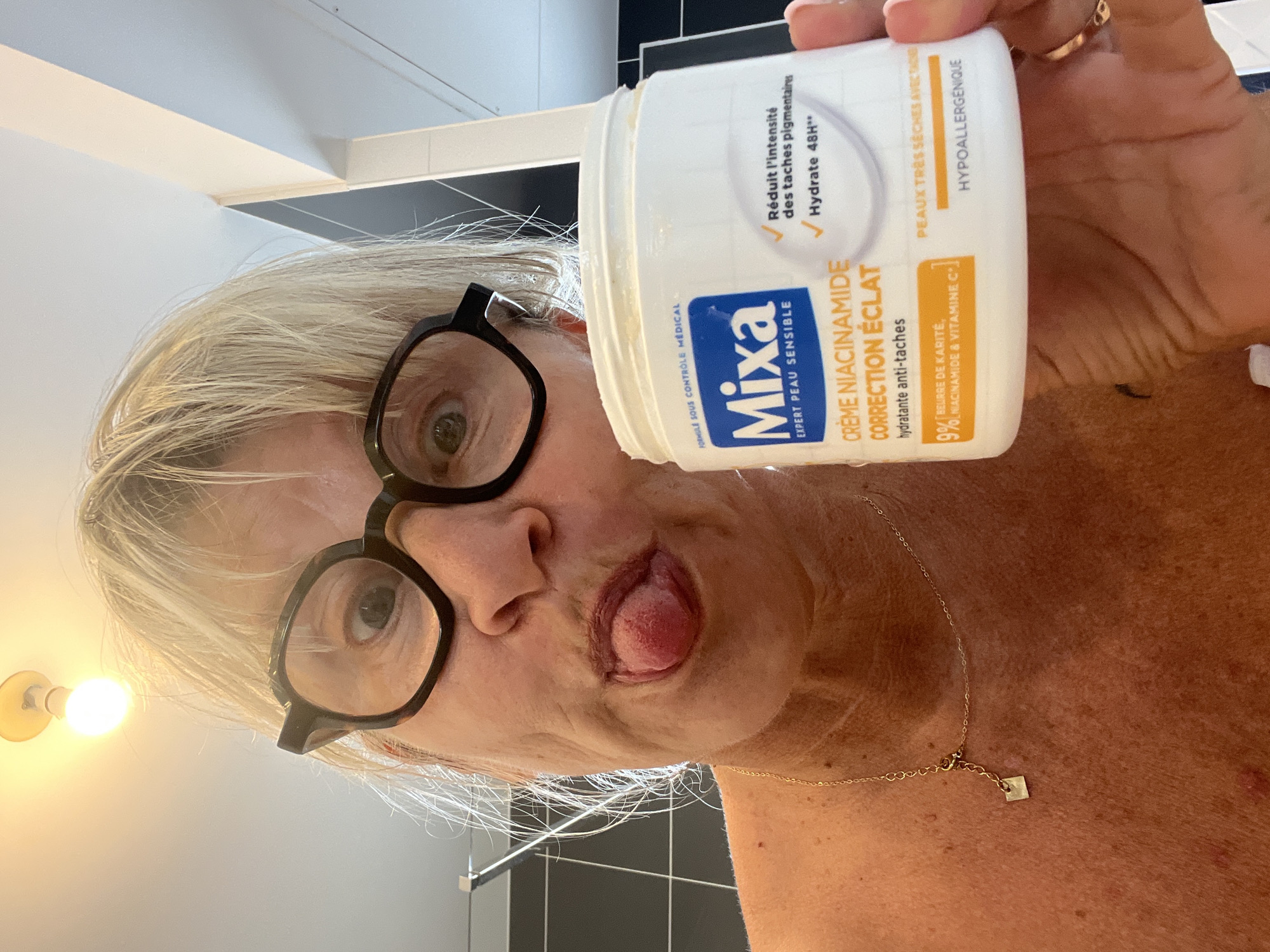 The image shows a woman, likely in her late 50s or 60s, with blonde hair, wearing large black-rimmed glasses. She is sticking her tongue out towards the camera and holding a white jar of "Mixa Creme Niacinamide Correction Eclat" in her right hand. The jar has a blue and white label with text in French. The woman is wearing a thin gold necklace with a small square pendant. The background appears to be a bathroom, with black tiled walls visible on the left. The lighting suggests an indoor setting, possibly a home bathroom. There is no indication of the city of Chambœuf, France, in the image.