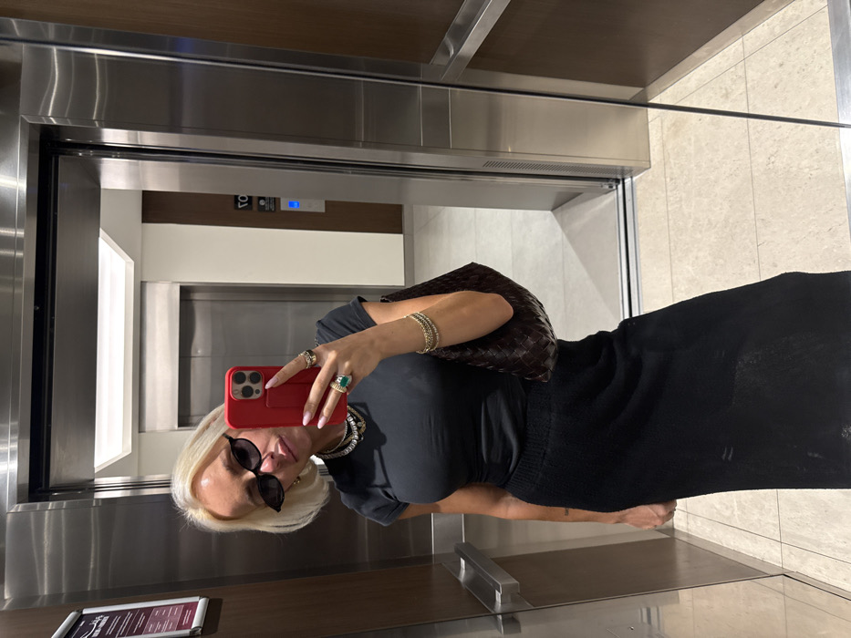 An adult female is depicted in a mirror selfie taken inside an elevator in Dubai, United Arab Emirates. The subject, with blonde, chin-length hair, wears small dark oval sunglasses, a black short-sleeved top, and a long black textured skirt. She holds a red smartphone in her right hand, which displays pink nail polish and multiple rings, including one with a large green stone. Several layered necklaces and thin bracelets on her left wrist are visible. A dark brown woven leather shoulder bag is carried on her left arm. The elevator interior features brushed stainless steel walls, a darker wood-paneled wall on the right, and a light-colored tiled floor. A horizontal handrail is present on the left wall. A framed sign with Arabic and English text is affixed to the left stainless steel panel. The reflection in the elevator's mirror shows a glimpse of an exterior hallway with light-colored walls, a matching tiled floor, another stainless steel elevator door, a vertical wooden accent panel, and a blue indicator light.