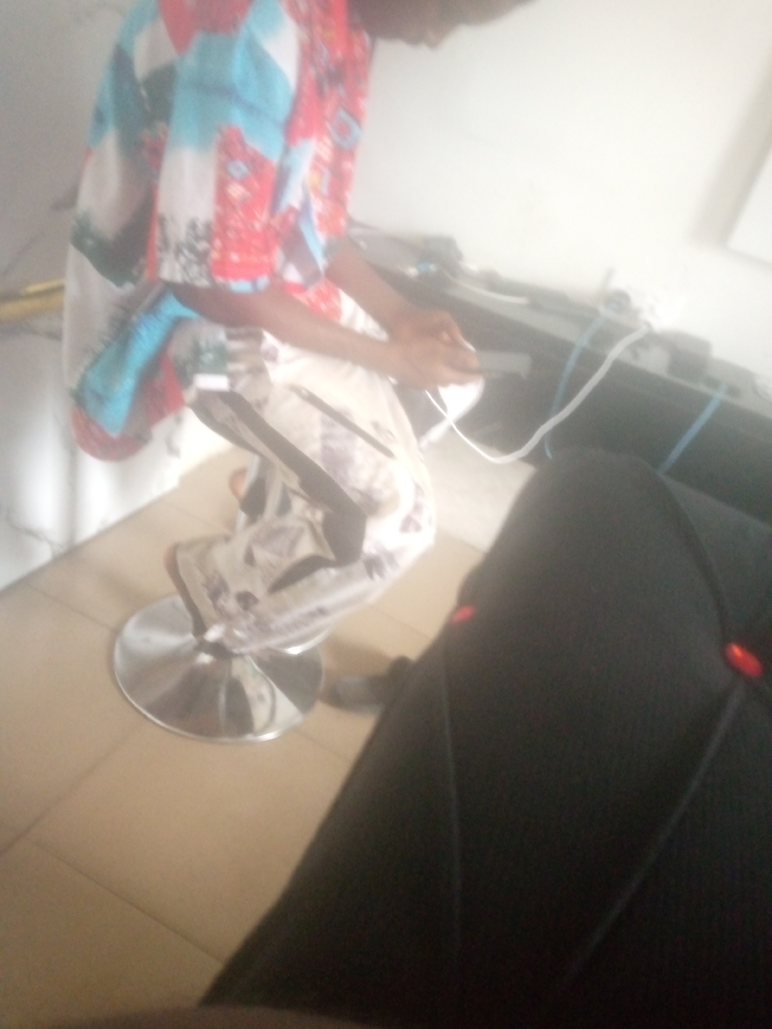 A person is seated on a chrome-legged stool in Jalingo, Nigeria. The person is wearing a colorful shirt with red, blue, and white patterns, and patterned pants. Their hands are holding a small, dark object, possibly a remote or phone, and they are looking down at it. The stool has a silver, circular base and is positioned on a tiled floor. In the background, there is a dark surface, possibly a desk or counter, with some wires and electronic equipment. The overall lighting suggests an indoor environment with a bright light source, potentially from a window or overhead lighting, creating some overexposure in parts of the image.