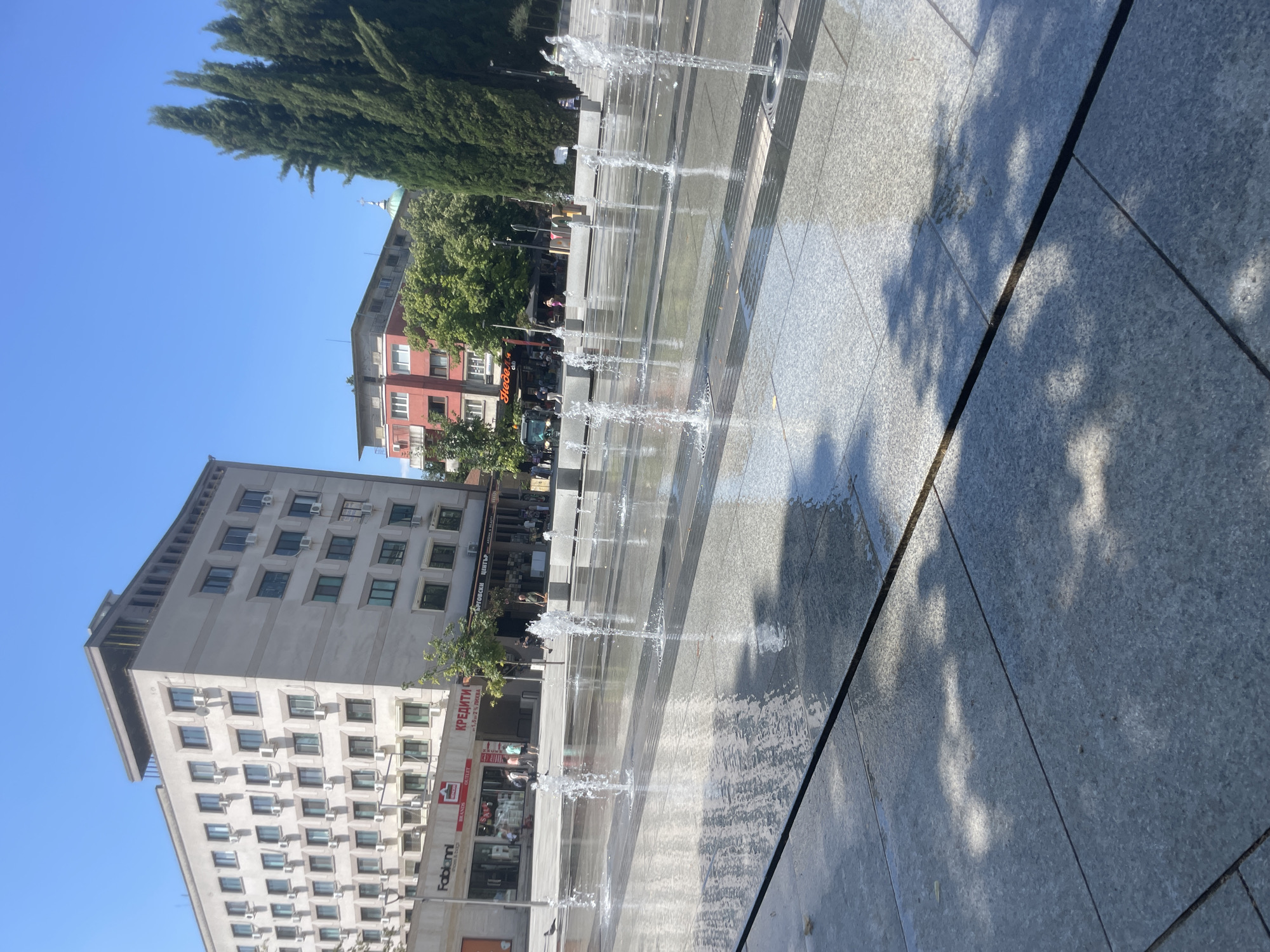 The image captures a sunny day in a city square with a water fountain in the foreground. The fountain features multiple jets of water spraying upwards from the paved ground, creating a shimmering effect on the wet surface. The square is paved with large, grey tiles. In the background, there are several buildings. To the left, a tall, light-colored building with many windows and air conditioning units is visible. Below it, a smaller building houses shops, including "Fabianni" and "КРЕДИТИ". Further to the right, a red building with multiple floors and balconies stands, surrounded by lush green trees. Tall, dark green cypress trees are prominent on the right side of the image. The sky is clear and blue, indicating good weather. There are a few people walking in the background, and a bird is visible near the fountain on the left. The overall scene suggests an urban public space, likely a pedestrian zone or a plaza designed for relaxation and recreation, located in the city of Stara Zagora, Bulgaria, based on the Cyrillic script on the buildings.