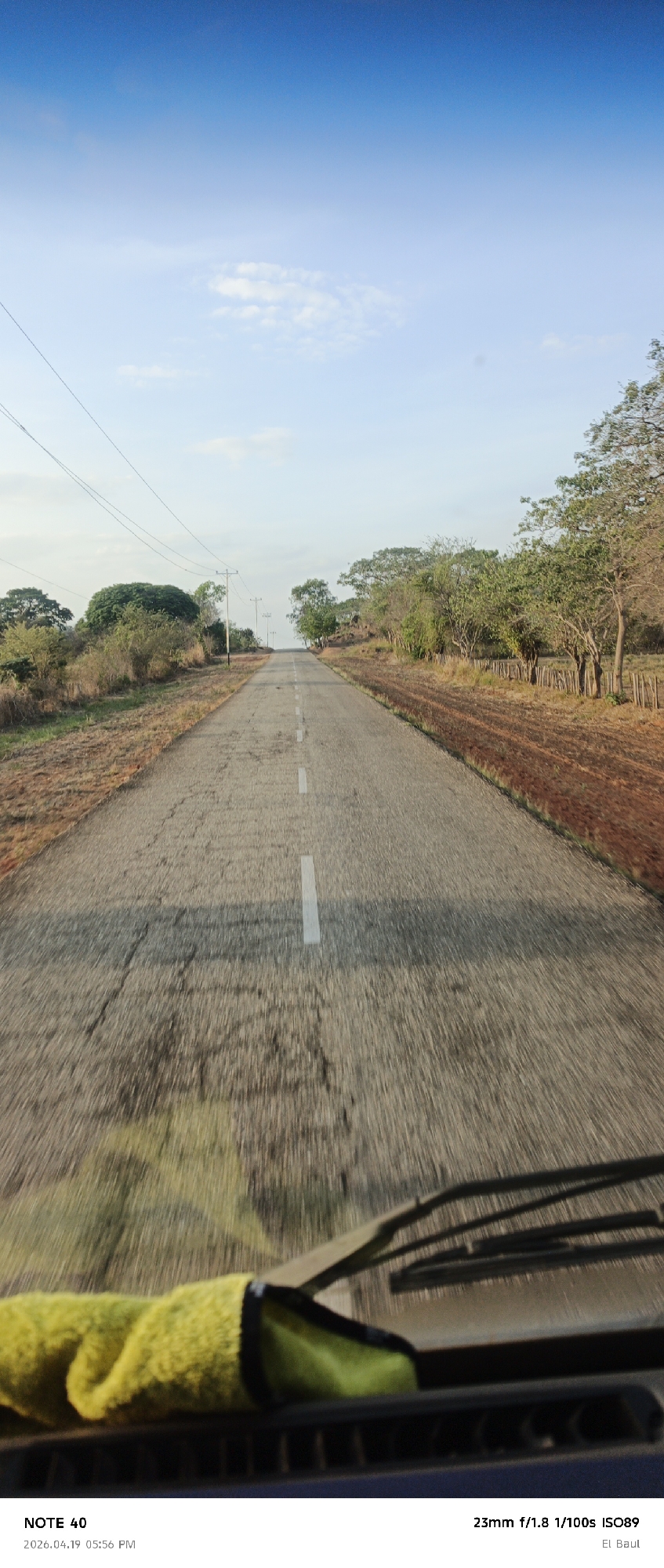 The image shows a long, straight, gray asphalt road with white dashed lines running down the center, presumably taken from the perspective of someone inside a vehicle. The road appears somewhat cracked and weathered. On either side of the road, there is dry, reddish-brown vegetation, transitioning into sparse trees and foliage. The sky above is a gradient, starting with a clear blue at the top and fading to a lighter, hazy blue near the horizon. Utility poles and power lines are visible in the distance along the roadside. In the foreground, parts of the vehicle's dashboard, a yellow and green cloth, and windshield wipers are visible, indicating the shot was taken from inside a moving car. According to the image's data, the photo was taken on 2026.04.19 at 05:56 PM, with a 23mm f/1.8 lens, at 1/100s, ISO89, in "El Baul", which is in La Cruz, Venezuela. There is no indication of the presence of people, subjects, actions, or interactions in the scene.