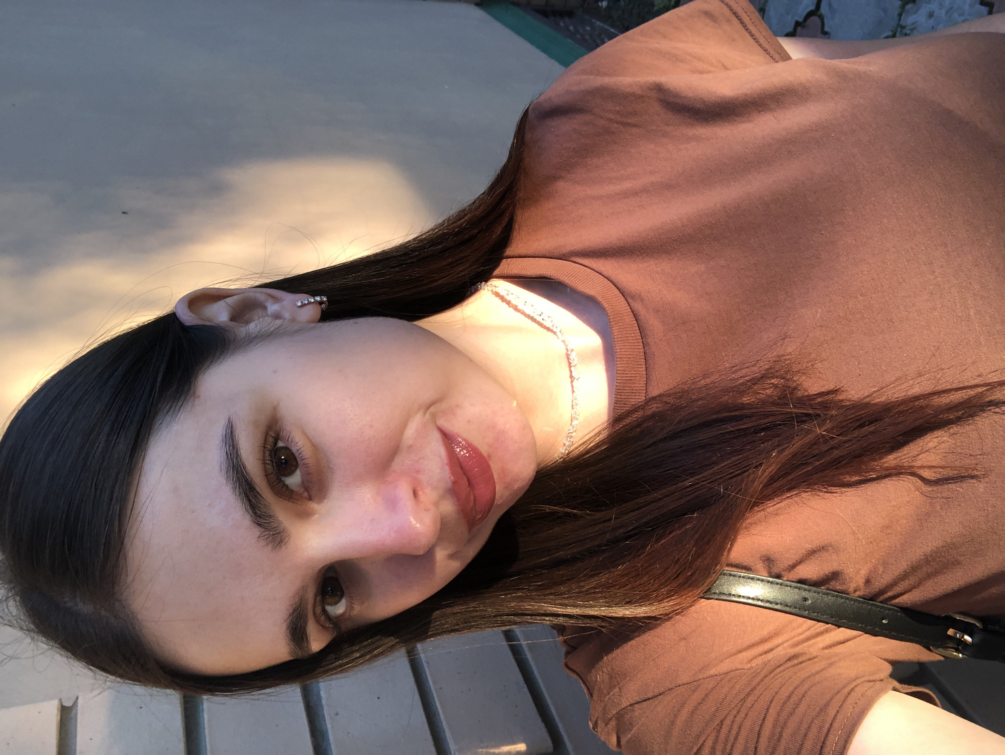 The media file is a portrait of a young woman, taken outdoors. She has long, dark hair and is wearing a brown t-shirt. A thin, sparkling necklace is visible around her neck. She has an ear piercing with multiple small, clear stones. The lighting suggests it is late afternoon, with warm, golden sunlight casting shadows and highlighting her features. The background is blurred, but appears to be a paved outdoor area with some textured elements, possibly a patio or walkway. The woman is looking towards the camera with a slight smile and her lips are glossy. The image is oriented sideways. There is no visible text in the media file. Given the provided location context, this scene could be in Chaikovsky, Russia.