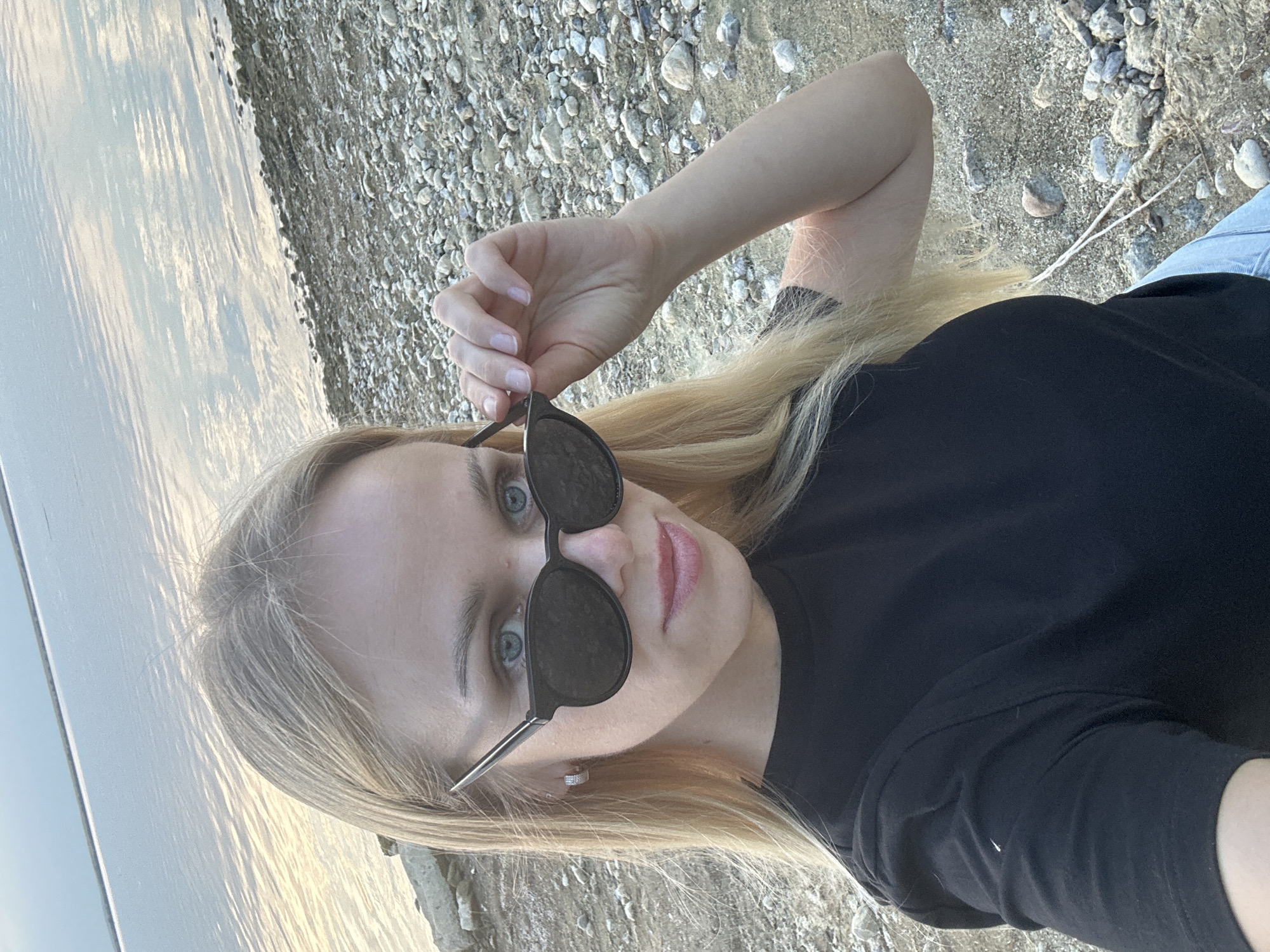 A blonde woman wearing sunglasses and a black t-shirt stands on a rocky shore, her hand touching the edge of her sunglasses. The background shows a body of water with a reflective surface under an overcast sky. The scene is located in Energetik, Russia.