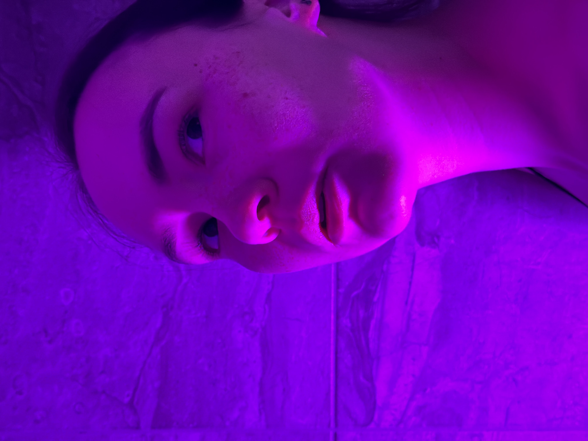 A close-up, waist-up, eye-level shot shows a young Caucasian woman with light brown hair, facing left. She is illuminated by purple light, which casts strong shadows and highlights on her face, revealing small blemishes. Her eyes are looking up and to the right, and her lips are slightly parted. A patterned wall, also bathed in purple light, forms the background. The image was taken in Chaikovsky, Russia.
