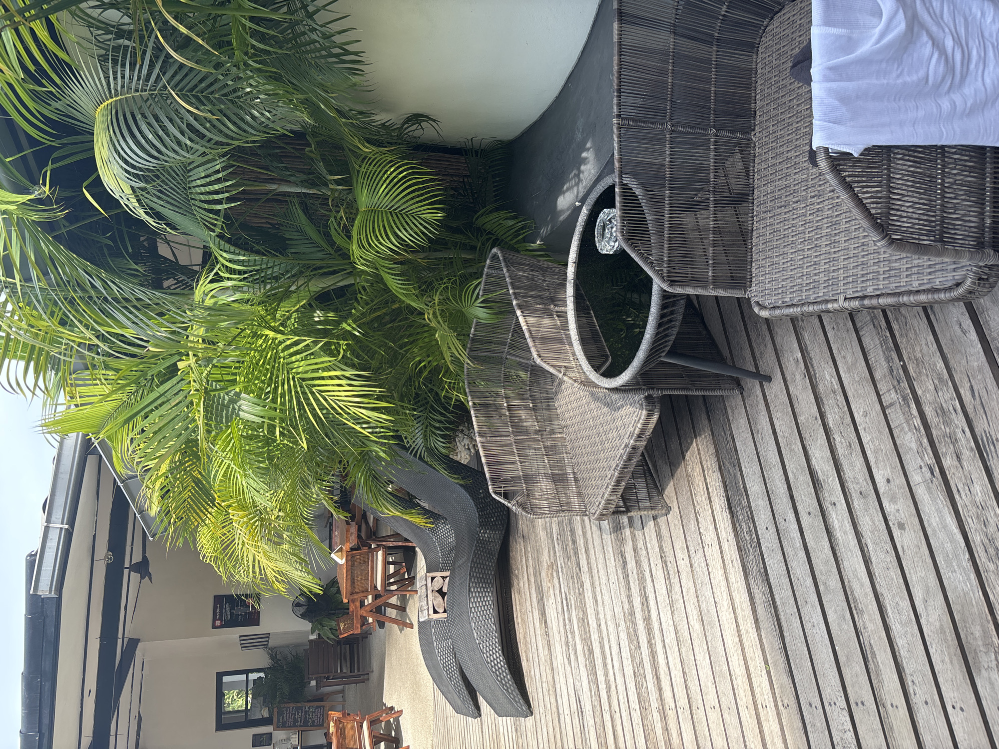 The image depicts an outdoor seating area with lush tropical foliage. Two dark, curved lounge chairs are placed on a weathered wooden deck. In the foreground, two wicker chairs and a circular glass-topped table with an ashtray are arranged on the deck. A white towel is draped over the arm of one of the wicker chairs. In the background, a row of wooden tables and chairs are visible, suggesting a dining or cafe area, with some palm trees and other greenery framing the scene. The environment appears to be a resort or restaurant setting in a tropical location.