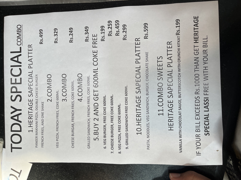 The image shows a menu board, likely from a restaurant in Sahibzada Ajit Singh Nagar, India. It lists various food combos and platters with their prices. The menu features several options, including a "Heritage Sapecial Platter" priced at Rs.499, a "Combo" with veg pizza, french fries, and coke for Rs. 329, another "Combo" with cheese burger, french fries, and coke for Rs.249, a "Combo" with grilled sandwich, french fries, and coke for Rs.349. There's also a "Buy 2 and Get 600ml Coke Free" option, followed by items 6 through 9, each offering a specific food item with a free 600ml coke. The menu further offers a second "Heritage Sapecial Platter" for Rs. 599, featuring pasta, noodles, veg sandwich, burger, and a chocolate shake. Lastly, there's a "Combo Sweets Heritage Sapecial Platter" for Rs. 199, which includes vanilla with chocolate sauce, butterscotch, and crunchy KitKat. The menu concludes with a special offer stating that if the bill exceeds Rs. 1000, customers will receive a free "Special Lassi" with their bill.