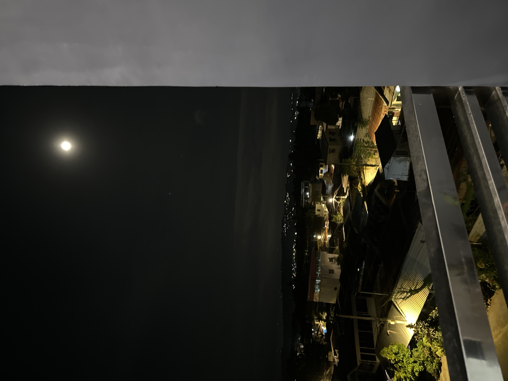 A nighttime scene from a high vantage point, possibly a balcony or rooftop in Davao City, Philippines. The frame's upper portion is occupied by a dark ledge and gray sky, while the lower left shows a body of water under a dark sky with the moon. The right side features a cityscape, including houses, streetlights, and a metal railing in the foreground.