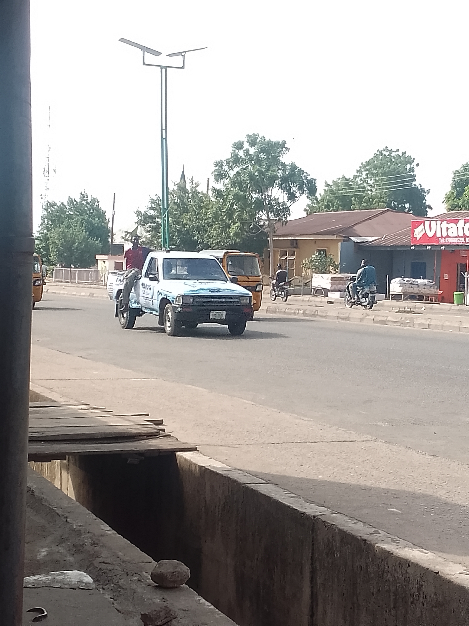This image depicts a daytime street scene in Pantame, Gombe, Nigeria. The perspective is from a slightly elevated position, looking across a paved road.
In the immediate foreground on the left, a dark, vertical support structure or pole is partially visible. Below it, several wooden planks cover a concrete drainage ditch, and a rounded rock rests on the ditch's edge.
The paved road occupies the mid-ground, featuring a concrete curb separating it from the foreground and a central concrete divider further down the street. Vehicles are in motion on the road. A light-colored pickup truck, possibly white with light blue graphic details including text that partially reads "TAX," is central, moving from right to left. A male individual in a red shirt stands upright in the bed of the truck, holding onto the cab. The driver is partially visible within the cab. A yellow tricycle (keke napep) is partially visible behind the foreground pole on the left, also moving left. Two individuals on motorcycles are visible on the right side of the road, moving away from the viewer. A portion of a yellow van or bus is visible on the far right, behind the pickup truck.
On the right side of the road, in the background, a series of single-story buildings are visible. One prominent building has a red facade with a white and red sign displaying "Vitafo" and partially legible contact information. A green refuse bin stands in front of this building. Adjacent to it is a building with a tan facade and a brown roof. Stacks of materials, possibly bags or building supplies, are positioned in front of these structures.
A tall, grey street light with two solar panels and two light fixtures extends upwards into the sky from the center-left of the mid-ground. Green trees are interspersed throughout the background and along the street. Overhead utility lines are visible against the bright, overcast sky in the upper right quadrant.