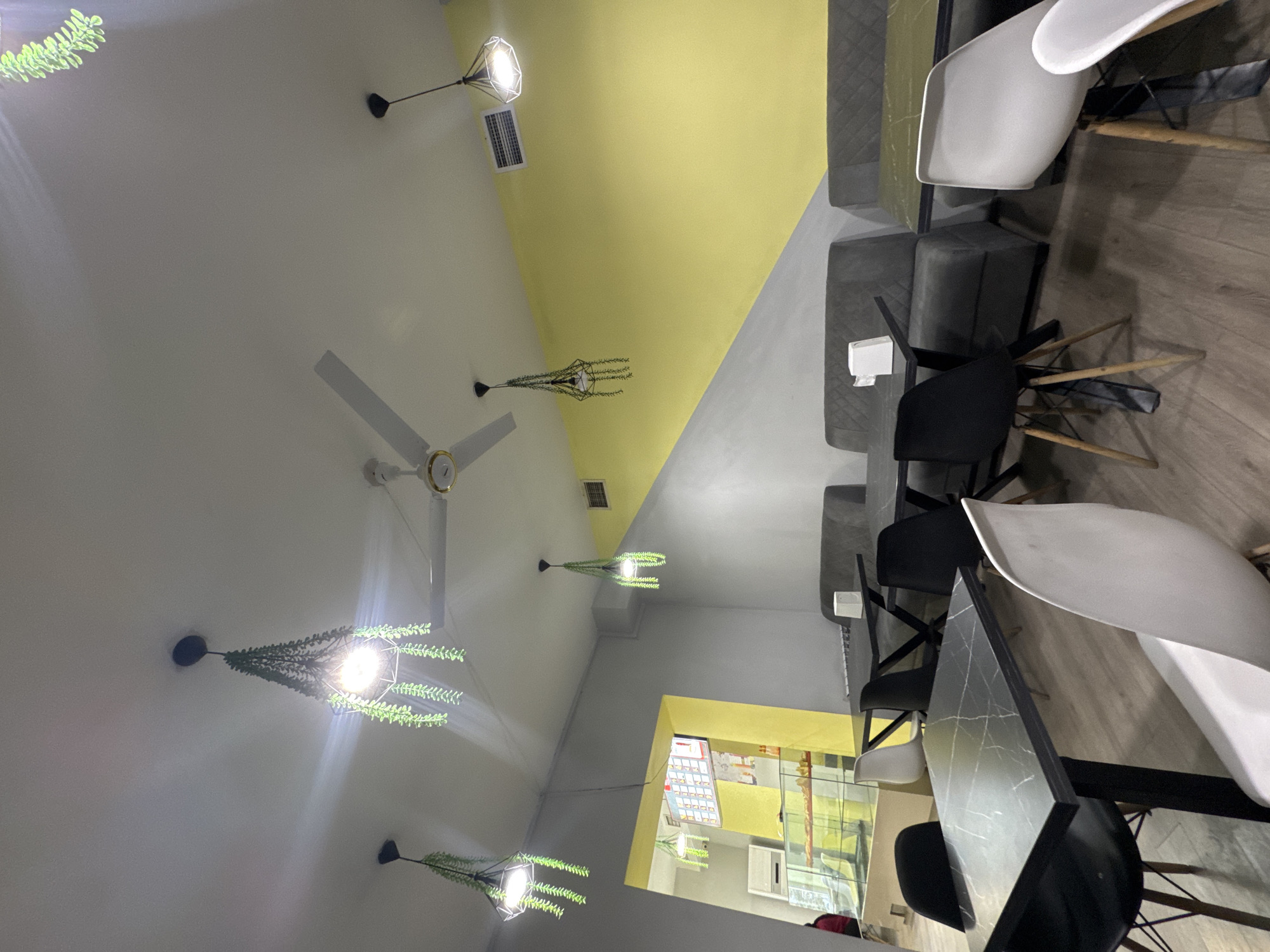 This media file captures the interior of what appears to be a modern cafe or restaurant, presented with a 90-degree clockwise rotation. The ceiling is a prominent feature, dynamically painted in sections of white, bright yellow, and light grey. Several decorative pendant lights hang from above, each enclosed in a geometric black frame and adorned with artificial green foliage, emitting a bright glow. Two white ceiling fans with gold accents are also mounted on the ceiling.
Seating arrangements include grey upholstered benches or sofas lining the walls, complemented by modern black and white chairs with wooden legs positioned around dark, possibly marbled, tables. The flooring is composed of wooden planks. In the background, a service or display counter with a glass case and shelving is visible, along with an air conditioning unit mounted on the wall. A brightly lit display board, resembling a menu or price list with columns of text, is also present, though the specific text is illegible due to blurriness.
No people are visible, and no specific activity is taking place, suggesting the image was captured when the establishment was quiet or closed. The ambiance is brightly lit by artificial light. The overall decor combines contemporary design with natural elements from the faux plants. This scene is located in Yerevan, Armenia.