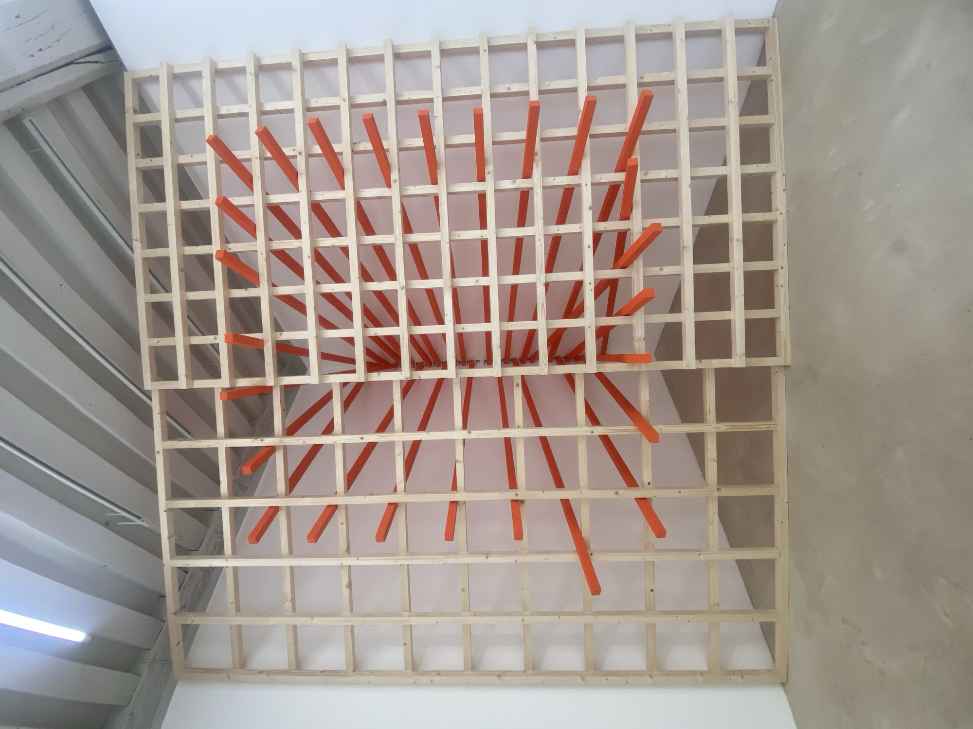 The image shows a close-up view of an art installation, likely situated on a ceiling. The installation consists of a wooden grid structure, seemingly made of unfinished light-colored wood, with uniformly spaced square openings. Interspersed throughout the grid are orange, rectangular beams of varying lengths that appear to protrude downwards from the ceiling. The beams are oriented radially, with a higher density near the center of the grid. The background is a plain white surface, presumably the ceiling of the room. The overall environment suggests an indoor space, possibly a gallery or exhibition hall. The image includes portions of adjacent walls or ceiling structures, one painted white and another with a textured, light-brown surface, potentially concrete or plaster. The context of the image is not in Paris.
