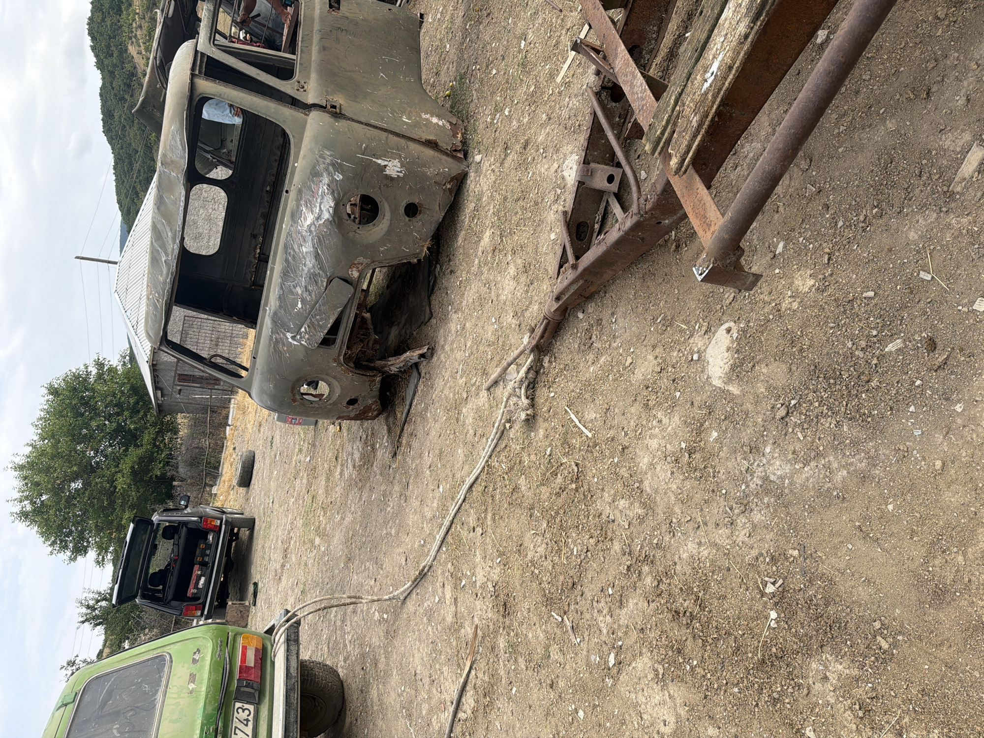 In Köhnə Quşçu, Azerbaijan, a rural setting features a collection of dilapidated vehicles and scrap metal. In the foreground, a rusty trailer frame lies on the dusty ground, with pieces of weathered wood attached. To the left, the rear of a green vehicle with license plate "743" is visible, with a thick rope extending from it across the ground. Behind it, a dark SUV with its trunk open sits on uneven terrain, displaying a toolbox inside. Further into the background, the corroded shell of a military-style vehicle, likely a UAZ, is prominently displayed, with its doors and roof missing. Another, similar vehicle chassis is partially visible behind it. The environment is a dirt lot with sparse vegetation and a few simple buildings in the background, indicative of a rural or semi-abandoned industrial area. The sky is overcast.
