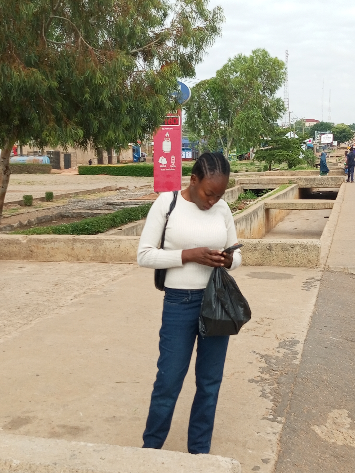 A woman stands on a concrete pathway, looking down at her mobile phone. She is wearing a white long-sleeved shirt and blue jeans. A black plastic bag hangs from her right hand. The background shows a street scene with trees, buildings, and a red signpost. In the distance, a blue truck and other vehicles are visible. The sky is overcast. The location appears to be the city of Tumfure, Nigeria, judging by the style of the buildings and the vegetation.