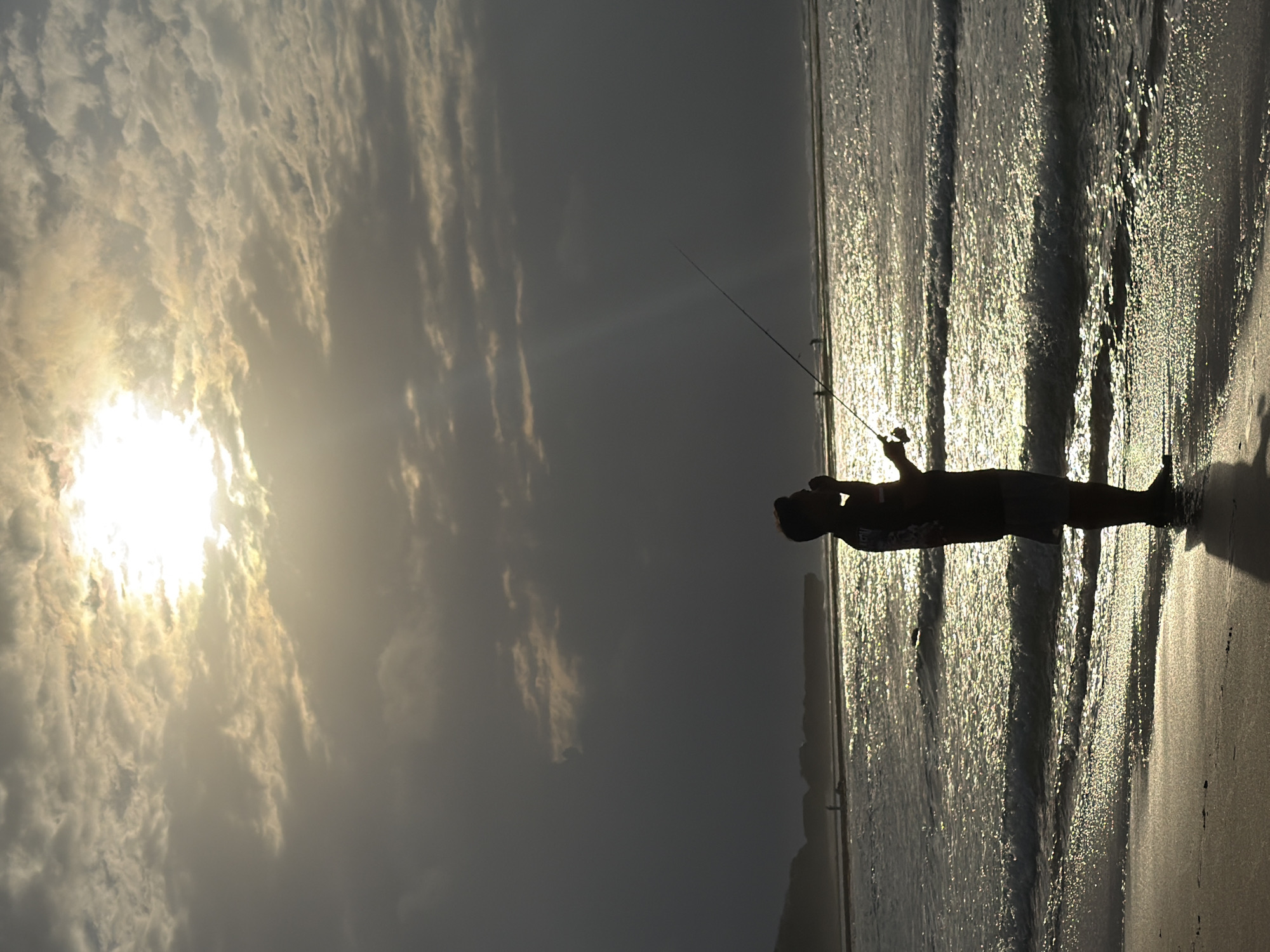 A silhouette of a person is standing on the beach in Kuta Selatan, Indonesia, holding a fishing rod. The person is facing the ocean, with the sun setting behind them, casting a bright glow across the sky and water. The waves are gently lapping at the shore, and the sand is wet and reflective. The sky is filled with dramatic clouds, some of which are illuminated by the sun, while others are dark and ominous. In the distance, there is a faint outline of the coastline, with a few other people visible on the beach. The overall mood of the image is peaceful and serene, with a sense of solitude and contemplation.