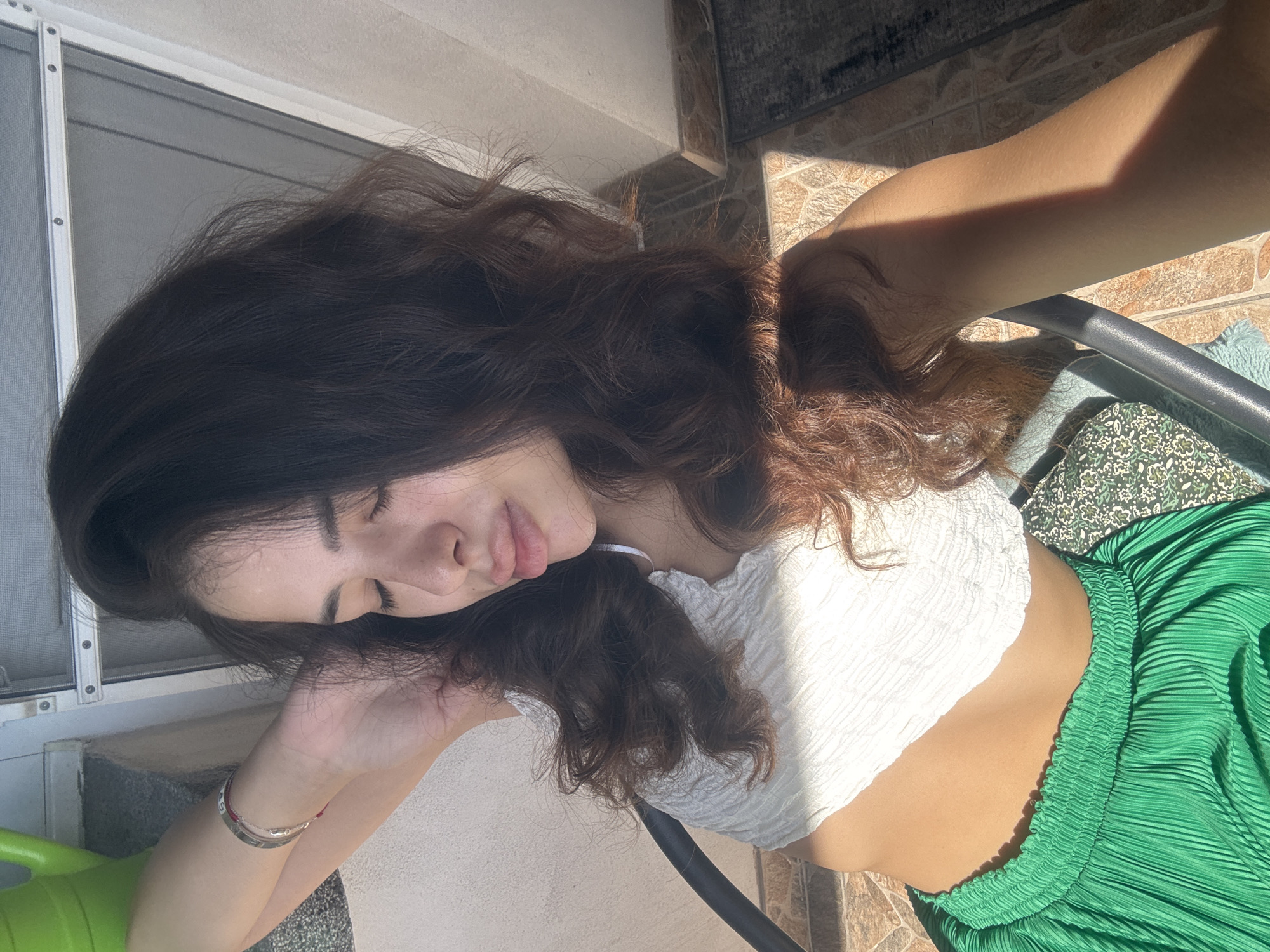 The image shows a young woman with dark, wavy hair and closed eyes, leaning back with her arm behind her head. She is wearing a white, textured crop top and a vibrant green pleated skirt. The lighting suggests she is outdoors, possibly on a patio or balcony, as indicated by the sunlight casting shadows and the presence of a chair. The background shows a tiled floor and what appears to be a window frame or door. There are no discernible landmarks or elements that would specifically place this scene in Plovdiv, Bulgaria.