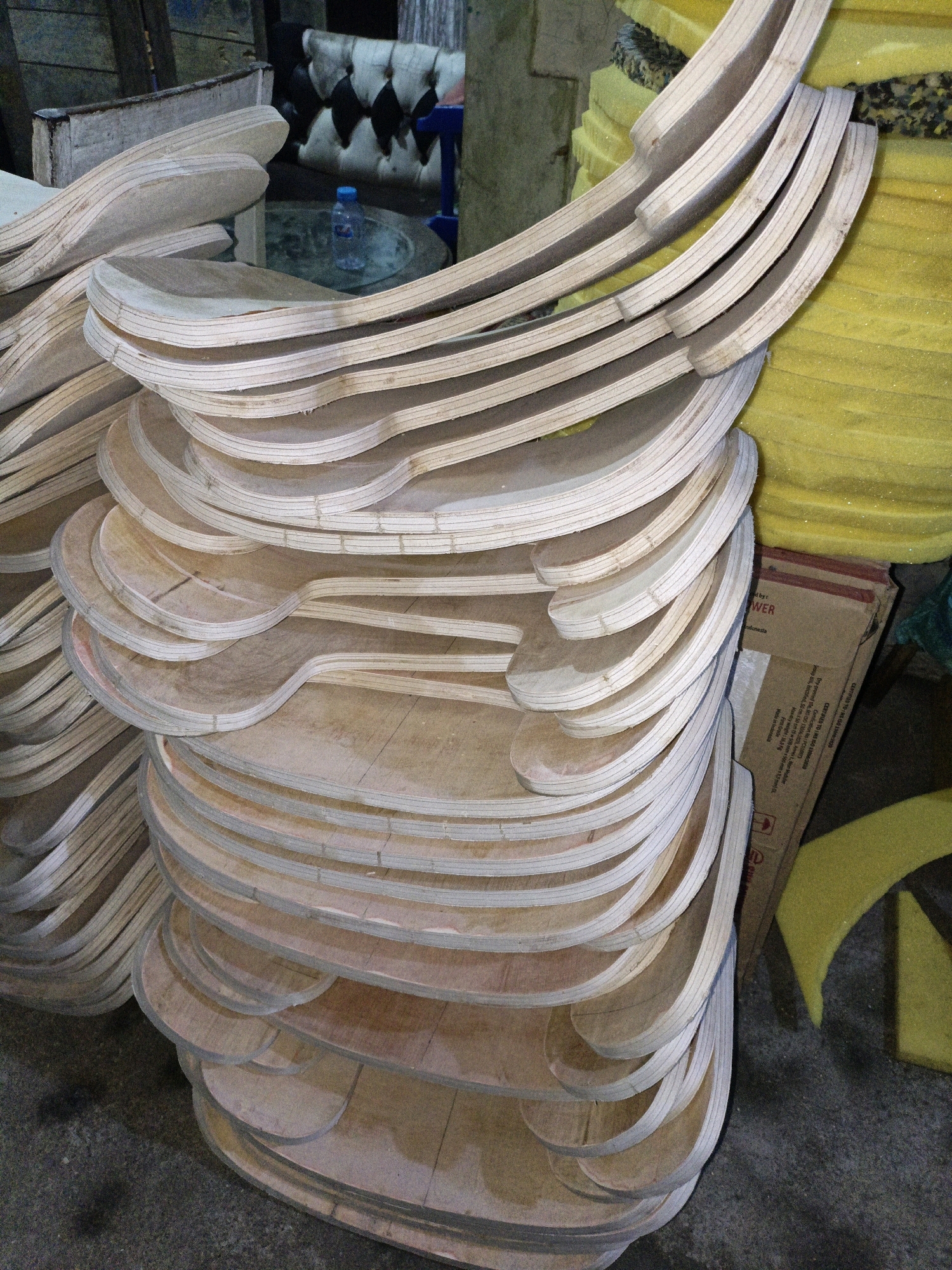 The image captures a detailed view of stacked, curved wooden elements, likely furniture components or parts of a larger structure, at a workshop or storage facility in Cikal, Indonesia. The wood pieces are predominantly light-colored, with visible wood grain and layered construction, suggesting they are made from plywood or similar laminated wood. Some pieces have a slightly pinkish or reddish hue on their edges, possibly from glue or treatment. The elements are neatly stacked in multiple vertical piles, creating a sense of organized storage and production.
In the background, a dark, tufted sofa is partially visible, along with a clear plastic water bottle on a circular glass-topped table, indicating a seating or break area within the facility. To the right, large sheets of bright yellow foam are stacked, and a brown cardboard box with printing, possibly shipping or product information, is also present. The floor is a rough, grey concrete surface, showing some dirt and debris. The overall environment appears to be a functional workspace, possibly a furniture manufacturing or repair shop, given the nature of the wooden components and the presence of materials like foam. No people are directly visible in the frame, but the organized arrangement of materials suggests ongoing work or recent activity. The lighting is natural and somewhat diffused, casting soft shadows.