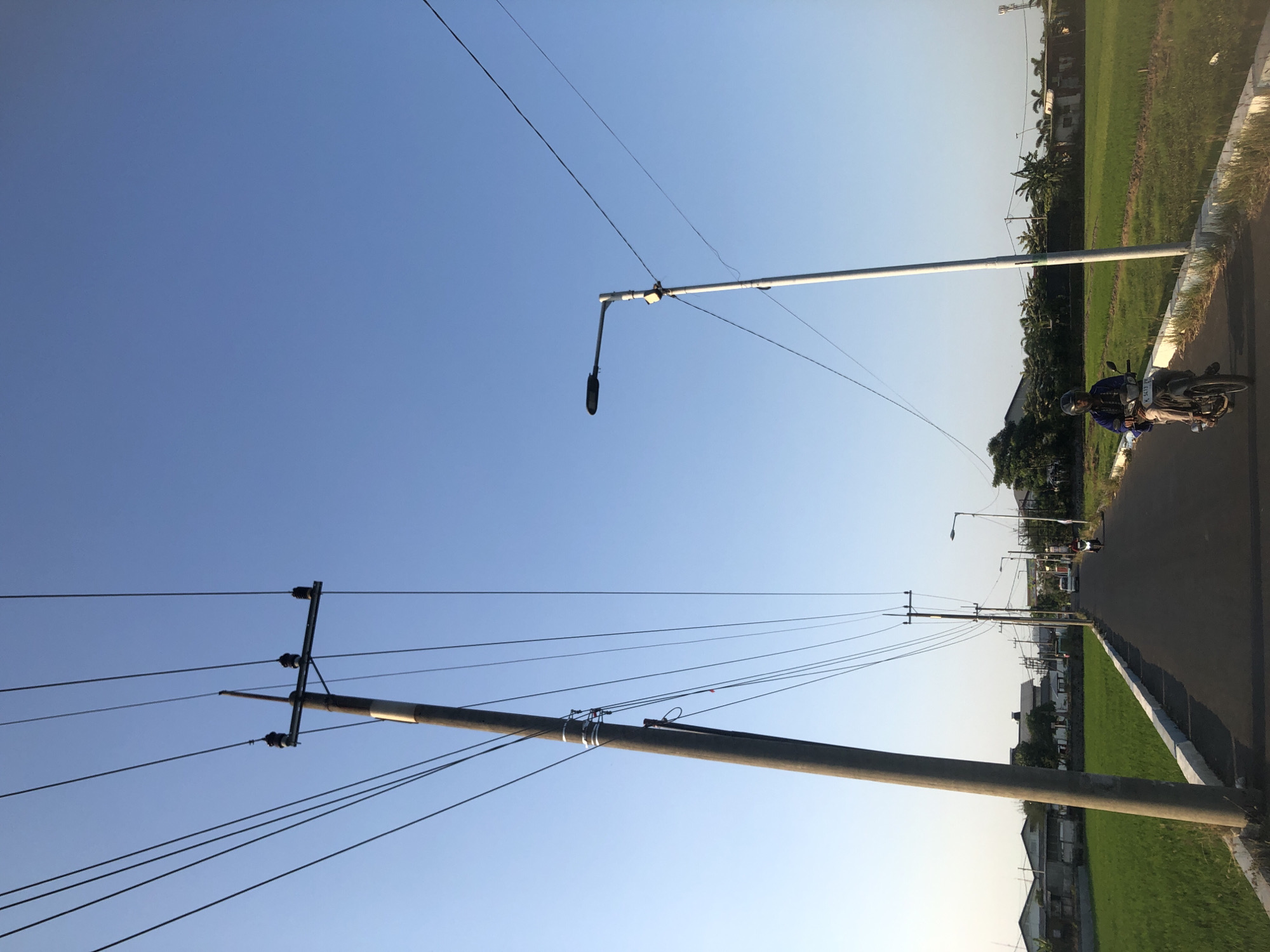 The image depicts a street scene in Kalitengah, Indonesia, under a clear blue sky. A paved road runs through the center of the frame, flanked by a lush green rice paddy on the left and more vegetation and buildings on the right. Several utility poles with overhead wires are visible along the road, with one prominent pole on the left side of the frame and another, a lamppost, on the right. A person is riding a motorcycle down the road, wearing a helmet and a blue jacket. The overall impression is one of a rural or semi-rural area during daylight hours.