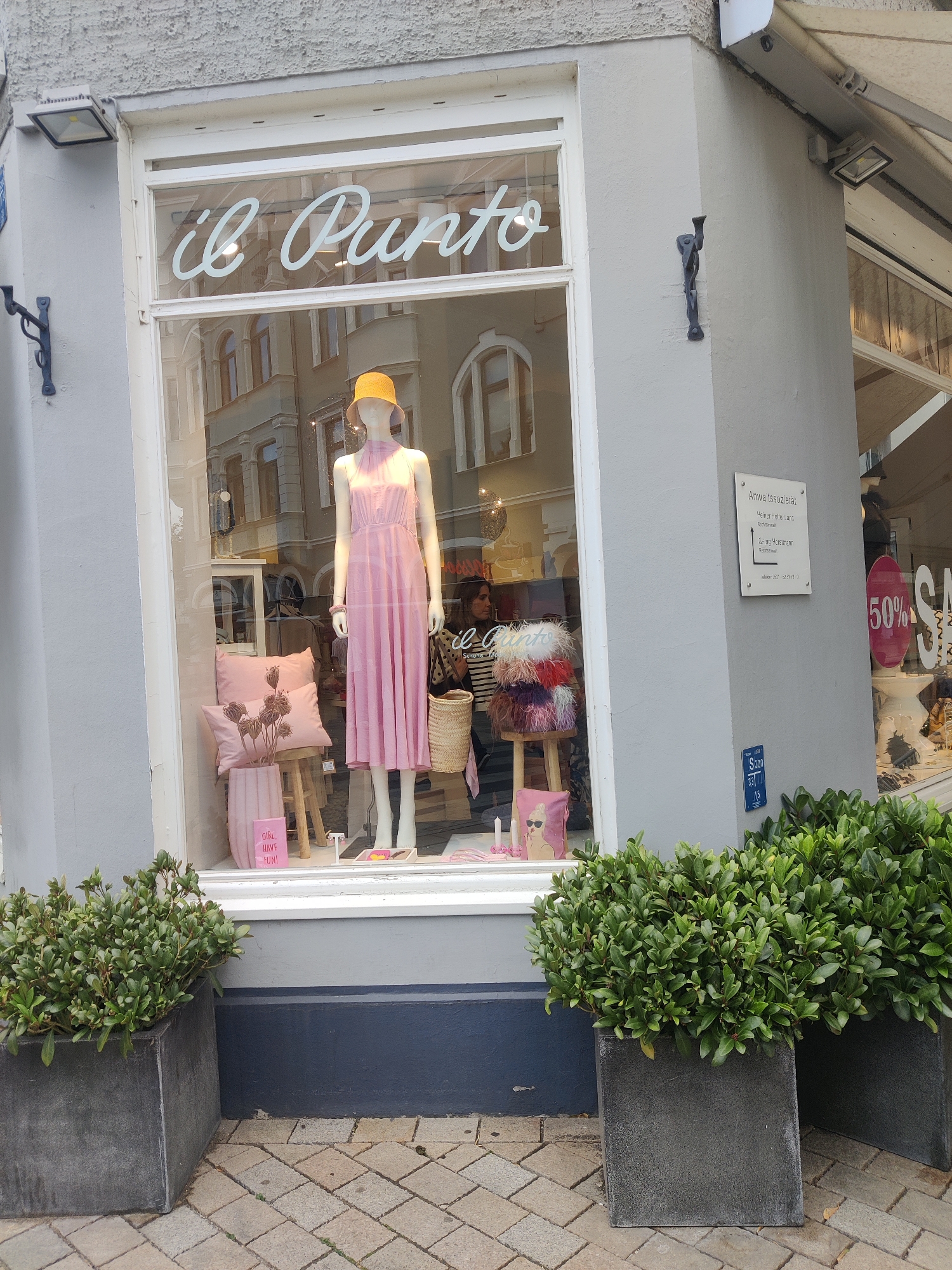 The image shows a storefront window displaying a mannequin wearing a pink dress and a yellow hat. The store is named "il Punto." In the window, there are also decorative pillows, a vase with dried flowers, and a woven basket. A sign on the wall next to the window reads "Anwaltssoziat" and other text, indicating a legal office. Another sign with "50%" is visible on the right side of the window, suggesting a sale. Outside the storefront, there are two large planters with green shrubs. The pavement is made of cobblestones. The scene appears to be a street in a city. Based on the provided information, this scene is located in Bielefeld, Germany.