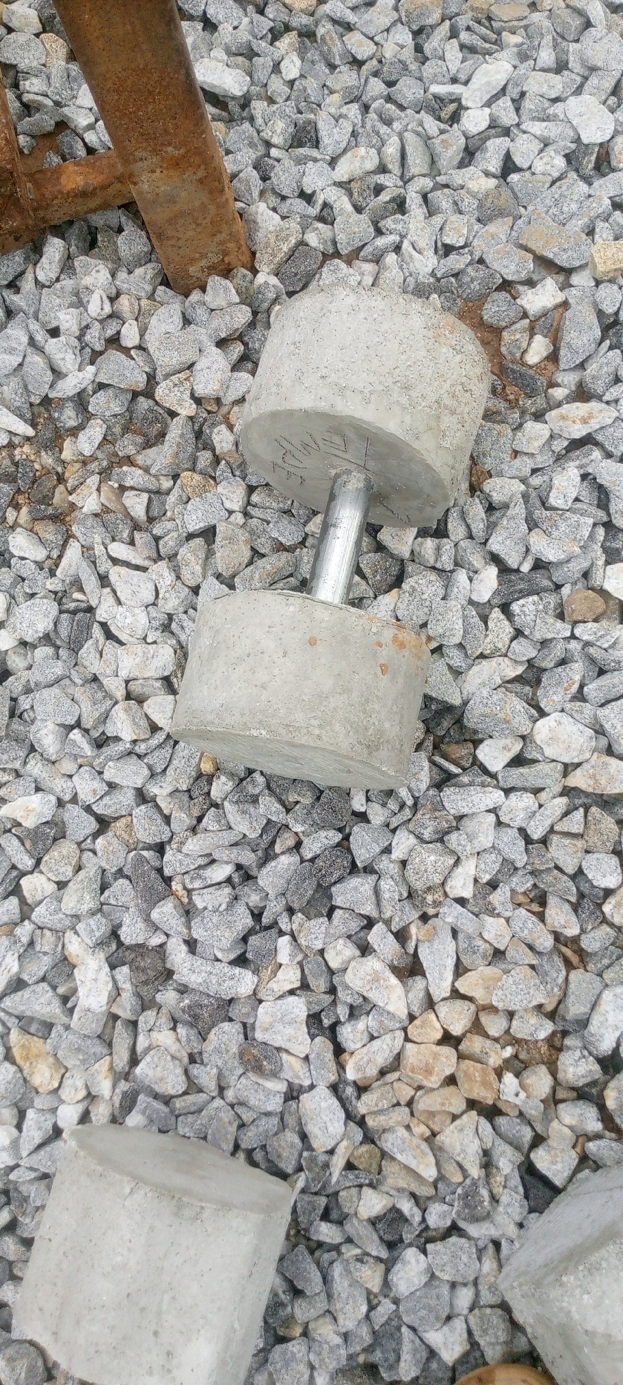 The image depicts a close-up, overhead view of a homemade dumbbell made from concrete and a metal bar, resting on a bed of gray, crushed stone. The dumbbell is positioned vertically, with its cylindrical concrete weights on either end of the metal handle. One of the concrete weights has some faint markings or engravings on its surface. In the background, a section of rusty metal, possibly part of a larger structure or equipment, is visible. The scene appears to be outdoors, likely in a construction or industrial area, given the presence of gravel and raw building materials. There are no people or animals in the image. The lighting is natural and somewhat diffused, suggesting an overcast day or shaded conditions. The overall setting is unrefined and functional, indicative of a site where construction or maintenance activities may be taking place.