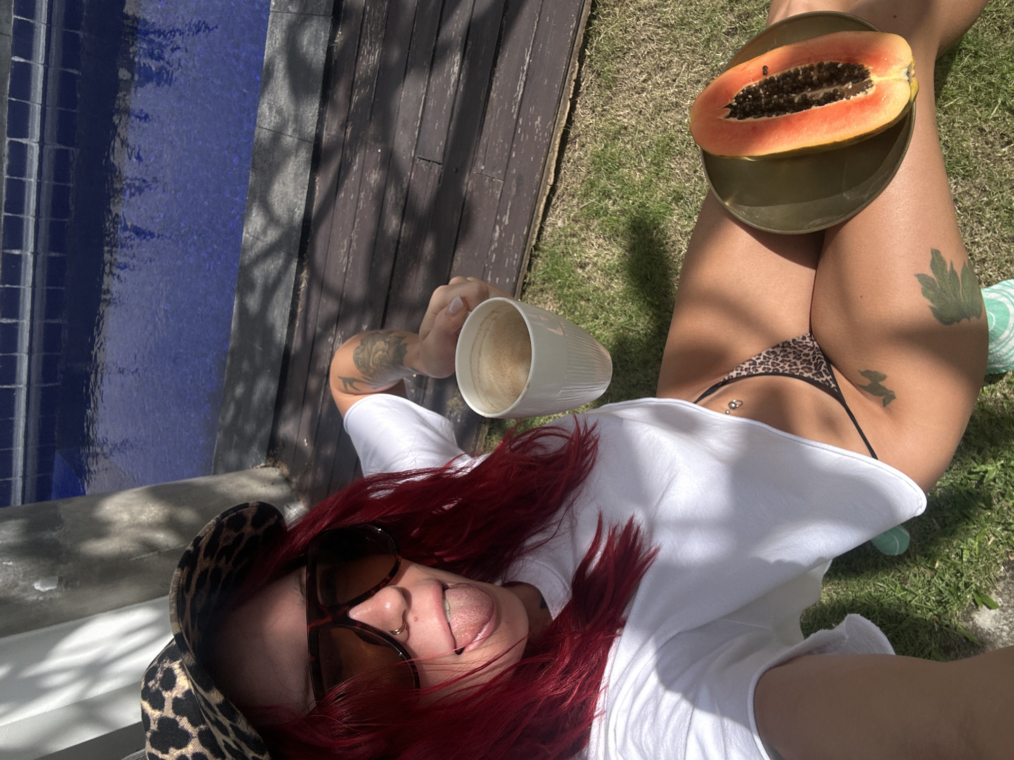 A woman with red hair and sunglasses is lying on the grass beside a pool and wooden deck. She is wearing a white shirt and a bikini. She is holding a white mug, and a halved papaya on a plate rests on her legs. The scene is outdoors, on a sunny day. A city cannot be determined.
