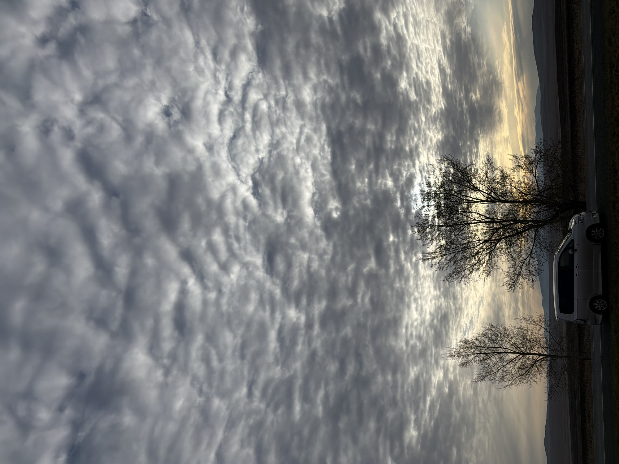 The image depicts a sky filled with layered gray clouds. The sky transitions into a landscape, where a car is parked next to a road, with bare trees nearby. Mountains are seen in the distance. The location could be the area around Первомайское, Russia, given the landscape.