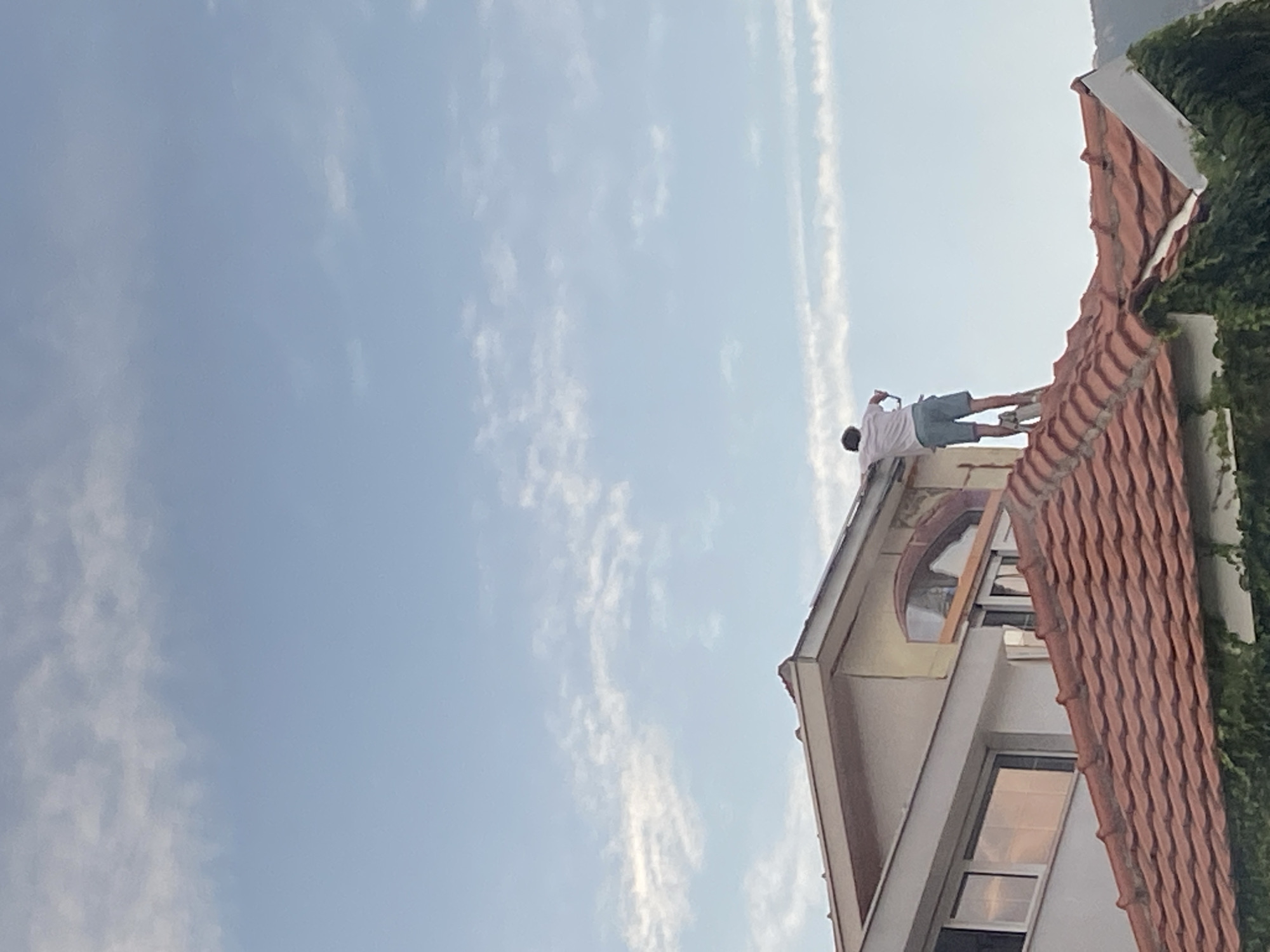 A person stands on the roof of a building in Budva, Montenegro. The building has orange tile roofing and a stucco exterior. A window with an arched top is visible. The sky is blue with scattered clouds. Green foliage grows on the side of the roof.