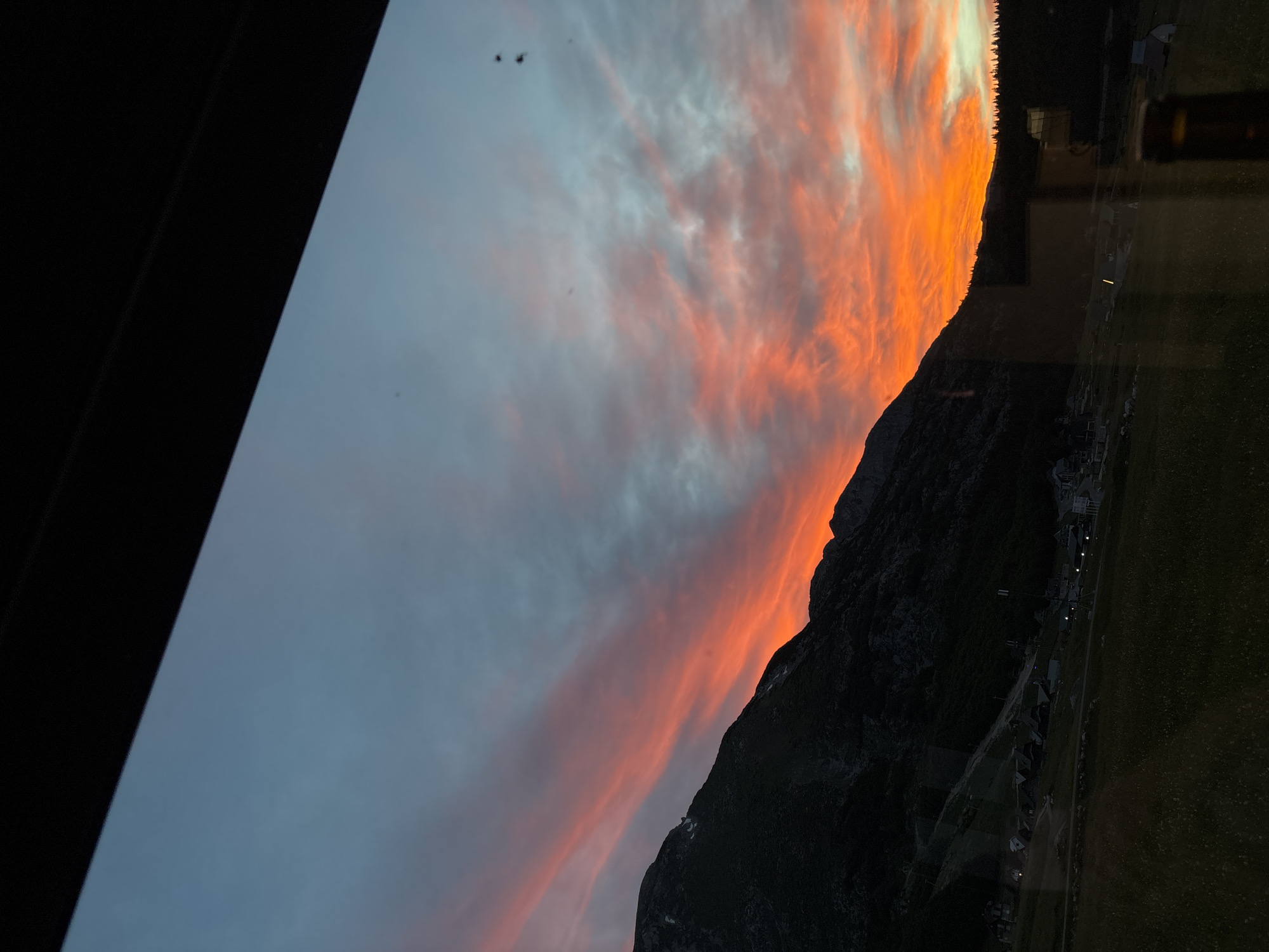 The image depicts a dramatic sunset over a mountainous landscape in Virak, Montenegro. The sky is ablaze with vibrant hues of orange, red, and pink, streaked with lighter blues and grays. Dark, silhouetted mountains dominate the left side of the frame, with a slope dotted with scattered buildings, suggesting a village or resort. The foreground is a dark, grassy field, with hints of vegetation and possibly some scattered wildflowers. The overall impression is one of a serene and picturesque natural environment at the end of the day.