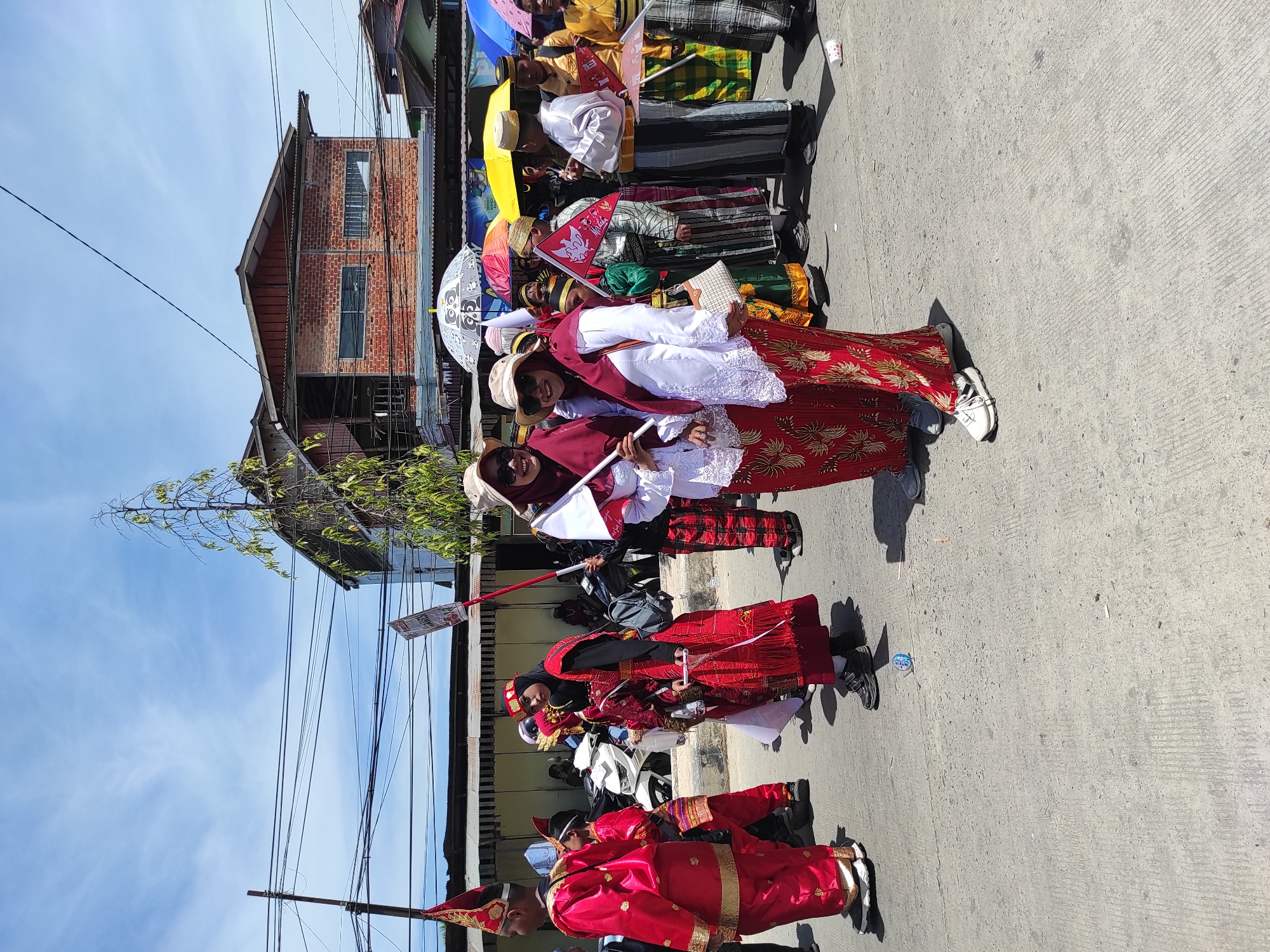 In Muara Jawa Ulu, Indonesia, a procession of people in traditional attire walks on a paved street under a clear, blue sky. The participants are dressed in vibrant red and gold garments, some with elaborate headdresses and others wearing headscarves. Many are holding flags and umbrellas. In the background, buildings with brick facades and corrugated metal roofs line the street, along with utility poles and tangled wires. The scene suggests a cultural event or celebration taking place outdoors during the daytime.