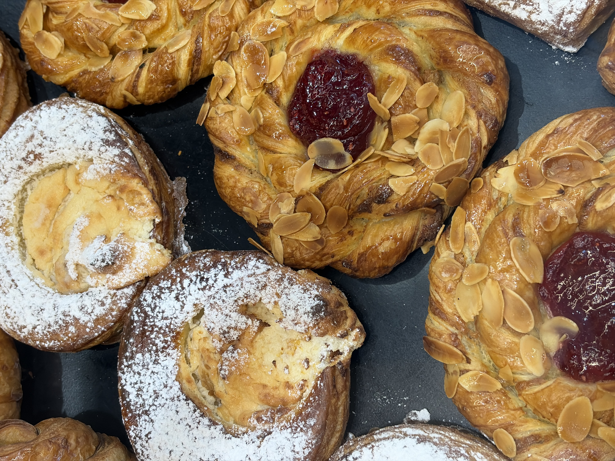 The image shows a close-up of various pastries on a dark, possibly slate, surface. There are at least two types of pastries: one dusted with powdered sugar and another type with a swirl design, topped with sliced almonds and a dollop of red jam. The pastries appear fresh and are likely for sale at a bakery or cafe. There is no information in the image to suggest this scene is located in Saint Petersburg, Russia.