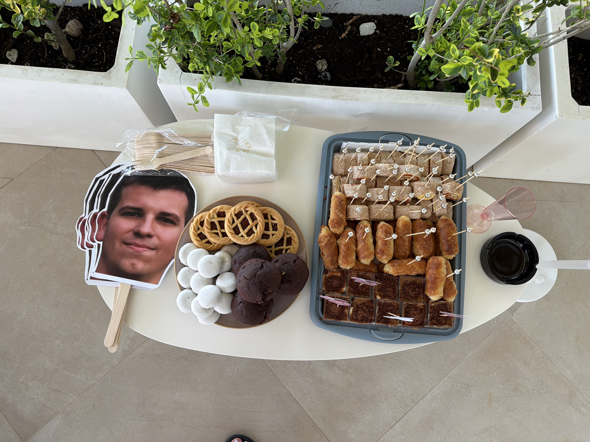 The image shows an outdoor table laden with a spread of snacks and a cardboard cutout of a man's face. A grey tray contains an assortment of small savory bites, including rolled sandwiches and what appear to be mini croissants, many of which are speared with decorative toothpicks. To the left of the tray is a brown plate with various sweet treats: small white powdered round pastries, dark chocolate cookies, and circular pastries with a lattice crust filled with jam. In the background, a stack of wooden disposable cutlery is visible, along with a packet of white napkins imprinted with gold lettering that reads "Team Bride". The table itself is round and white, set against a tiled floor. To the right of the table are white planters with lush green foliage. The overall setting suggests a casual gathering or party. The specific location in Kassandra Municipal Unit, Greece, is not determinable from the visual information provided.