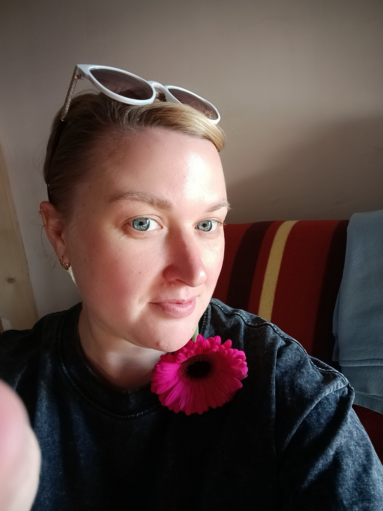 A woman is taking a selfie indoors. She has fair skin, short blonde hair, and blue eyes. White sunglasses rest on top of her head. She is wearing a dark-colored t-shirt, and a pink gerbera daisy is positioned near her neck. Behind her is a striped blanket in red, orange, and beige hues, as well as a blue blanket. The location is Chaikovsky, Russia.