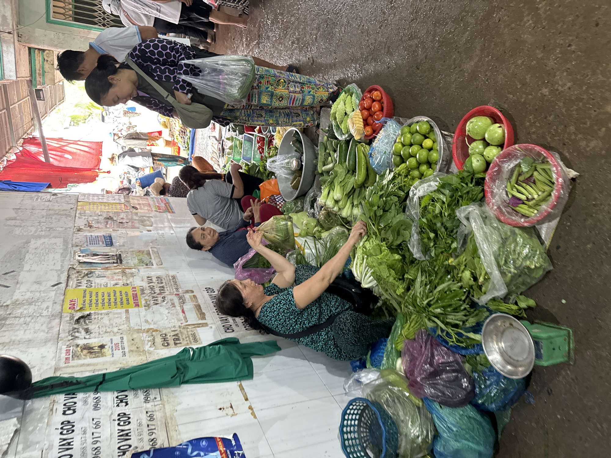 The image depicts a bustling outdoor market scene in Mũi Né, Vietnam. In the foreground, several women are arranging and selling a variety of fresh vegetables. One woman in a blue and green patterned outfit is crouched down, offering a plastic bag of vegetables to a customer. Another woman, wearing a black and purple patterned top, stands nearby with a green bag over her shoulder. A diverse assortment of produce is displayed, including leafy greens, cucumbers, tomatoes, limes, and okra, some of which are contained in plastic bags and baskets. A small green weighing scale is visible on the ground. In the background, more market stalls and people can be seen, with a covered walkway overhead. The ground is wet and appears to be concrete or pavement. The walls in the background are covered with posters and advertisements. The overall atmosphere is one of active commerce and local life.