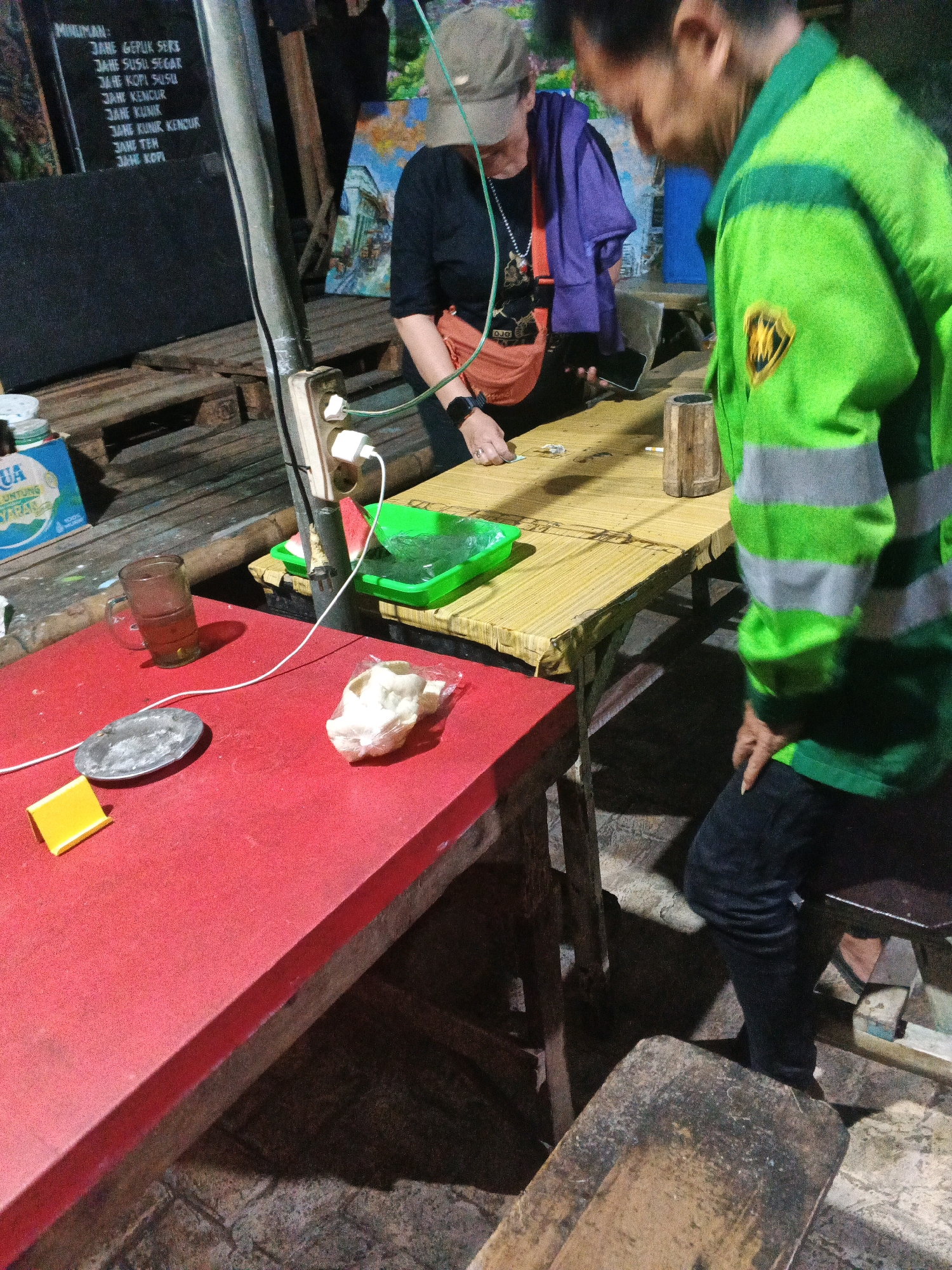 The image depicts an outdoor scene at night, likely a food stall or small restaurant, in Ungaran, Indonesia. Two individuals are present. One person, wearing a bright green jacket with reflective stripes and black pants, is standing and looking down. The other person, wearing a beige cap, black shirt, and an orange fanny pack, is seated and appears to be interacting with something on a bamboo table. A red table is in the foreground with a glass of tea, a small metal dish, a yellow object, and a plastic bag containing white food items. Various surfaces and objects suggest a rustic, informal setting. Electrical outlets are visible, with a wire connecting them to a device on the bamboo table. A menu is partially visible on a black board in the background. The overall lighting is dim, typical of nighttime street vendor environments.