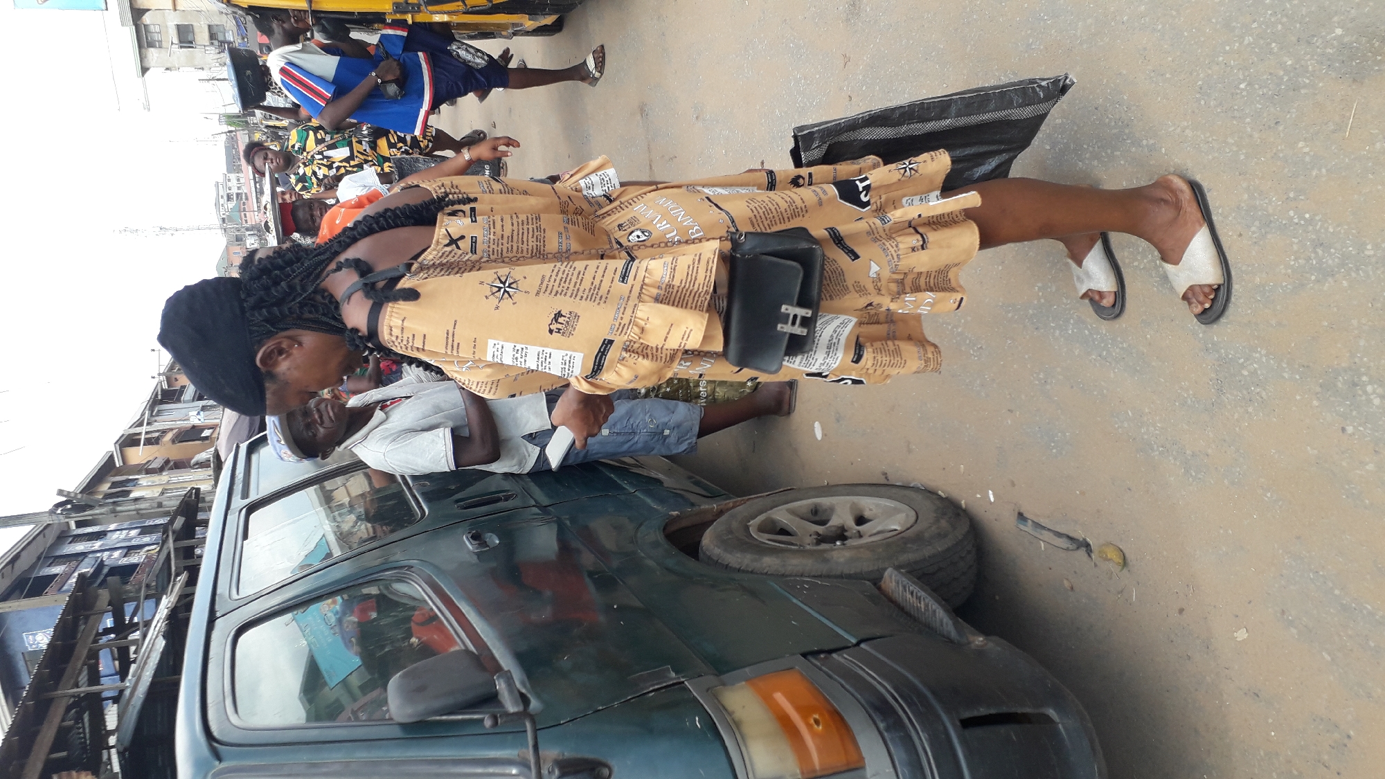 On a city street in Onitsha, Nigeria, a woman in a tan newspaper-print dress stands near a dark green van with a flat tire. She wears a black beret, has dark hair, carries a small black purse with an "H" on it, and wears white sandals. The van's passenger side and front corner are visible. A man in a white shirt and hat is near the van, possibly inspecting it. Other people are present in the background, including a man on a tricycle. Buildings can be seen in the background, suggesting an urban environment. The street surface appears to be paved but somewhat dusty or dirty.