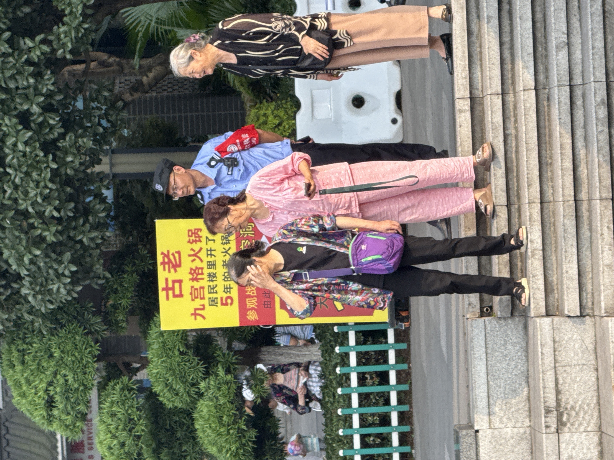 The image depicts a scene in Yuzhong District, China. Several people are standing on steps next to a yellow sign. The sign has Chinese characters, which translate to "Old Nine Palaces Hot Pot, Opened for 5 years in the residential building, Observation Battle". A woman wearing a black and white top and light brown pants stands at the top of the stairs. Below her, a man in a blue police uniform stands. Another woman in a pink jumpsuit stands next to him. A fourth woman in a black shirt, a colorful patterned jacket, and black pants with a purple bag over her shoulder is near the sign. A green fence is between the sign and the road with trees.