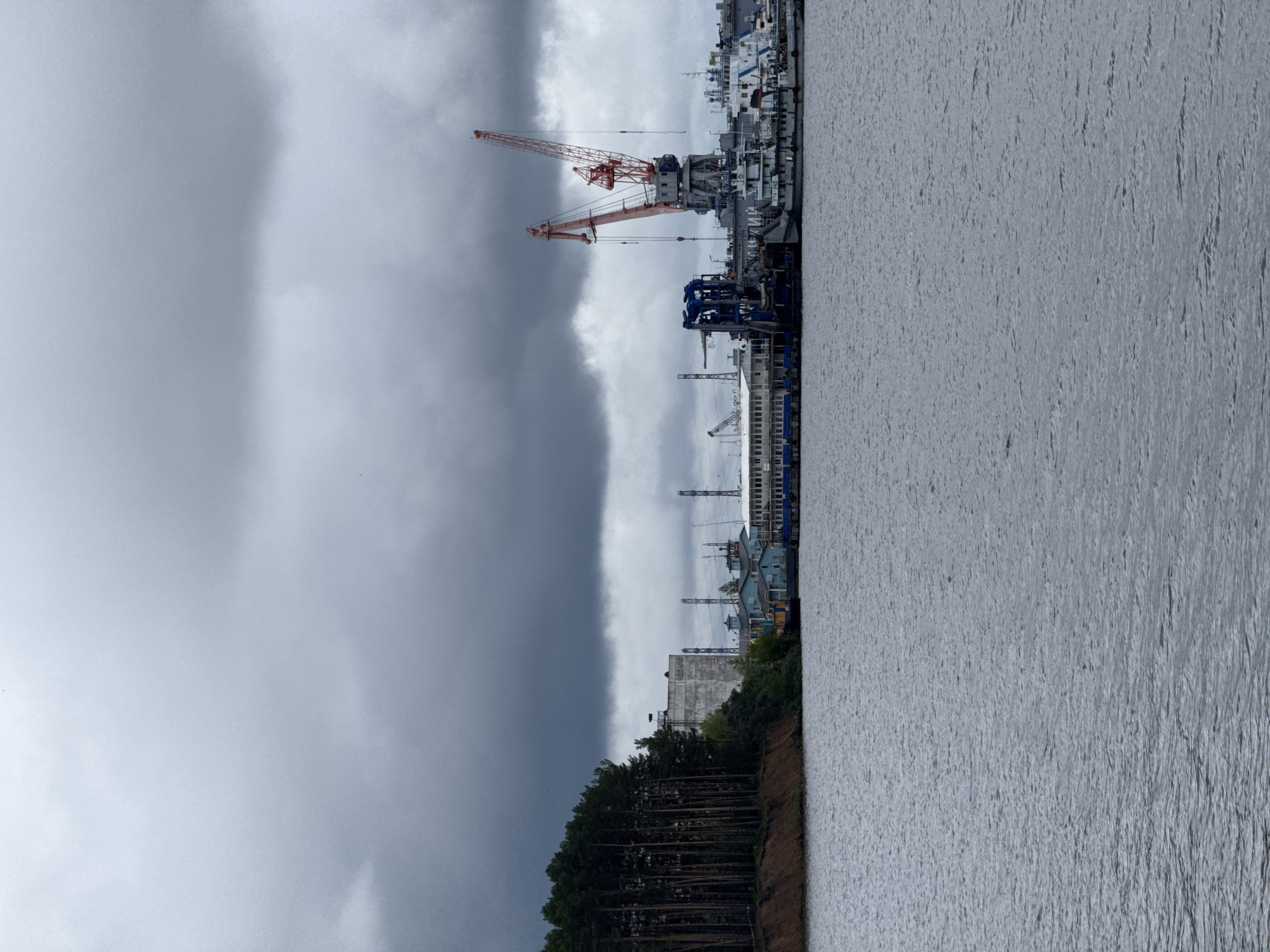 This image captures a wide view of an industrial waterfront in Chaikovsky, Russia, under an overcast sky. The upper two-fifths of the frame are filled with a dense, unbroken expanse of dark grey, turbulent clouds. The lower three-fifths of the image consist of a body of water, likely a river, exhibiting a textured surface with ripples and appearing dark grey.
On the left side of the midground, a steep bank of reddish-brown earth rises from the water, densely covered with a vertical line of tall, dark green coniferous trees extending into the upper midground. To the right of the treeline, across the water, the opposing shore reveals an industrial complex. A large, multi-story, rectangular concrete building with a flat roof is visible behind the trees. Extending further right along the waterfront are several smaller, light blue-colored buildings with white roofs.
A series of large industrial cranes dominates the center-right midground. A prominent blue gantry-style crane or similar heavy machinery is positioned closer to the left of the complex. To its right are two substantial red and white lattice boom cranes, one taller and more upright, the other slightly angled. Numerous power lines, poles, and other industrial structures are visible behind these facilities. Several vessels are docked along the waterfront: a grey workboat or utility vessel, identified with the text "OTA-942" on its hull, is moored beneath the red cranes. To its right, partially obscured, is a white-hulled vessel with blue detailing, possibly a patrol boat or similar craft. No human figures are discernible in the scene. The overall impression is that of an active port or shipbuilding facility under heavy cloud cover.